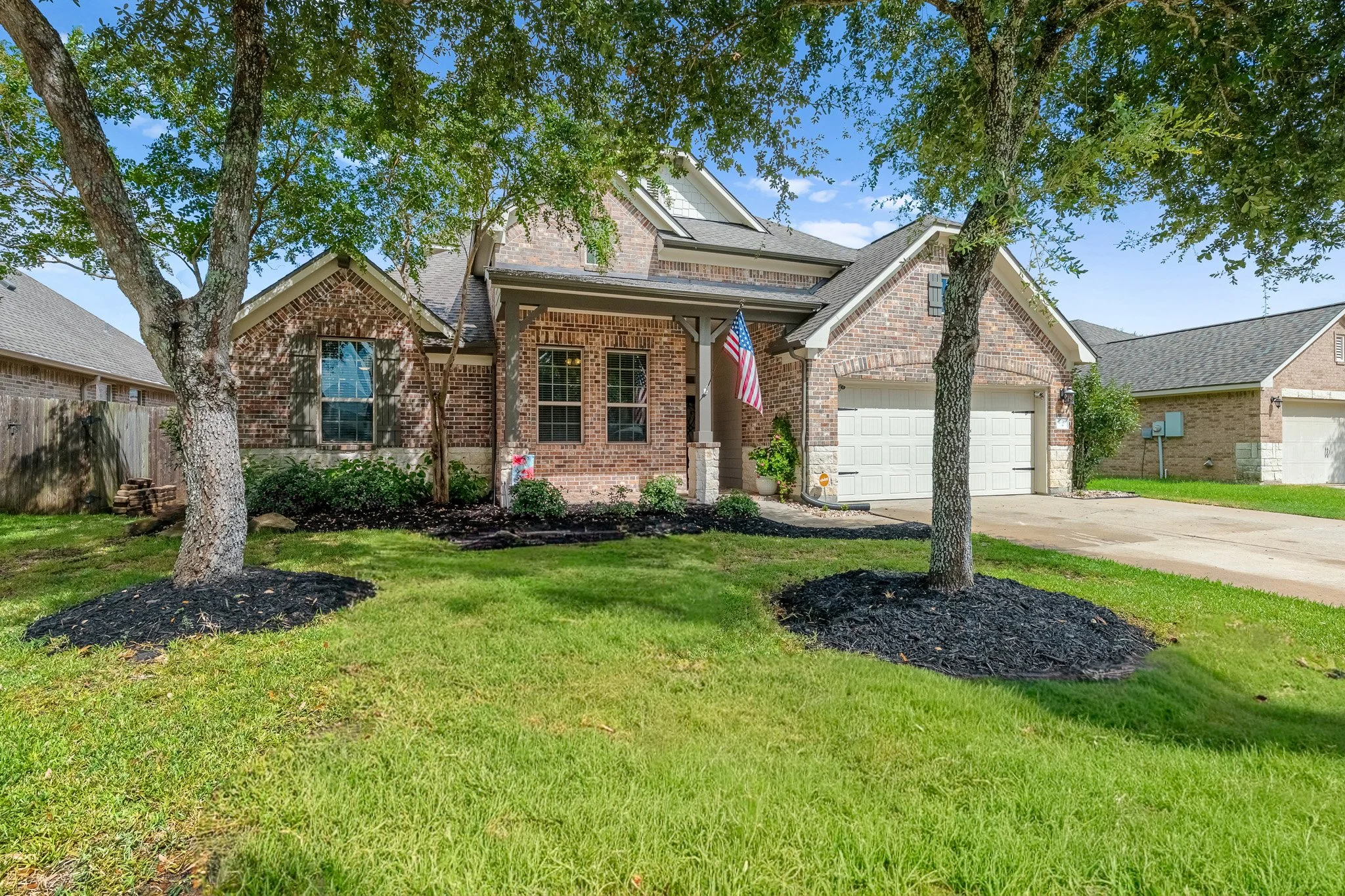 Front view of a brick house with a white garage door, two trees in the yard with mulch beds, and an American flag hanging near the porch in a sunny neighborhood.