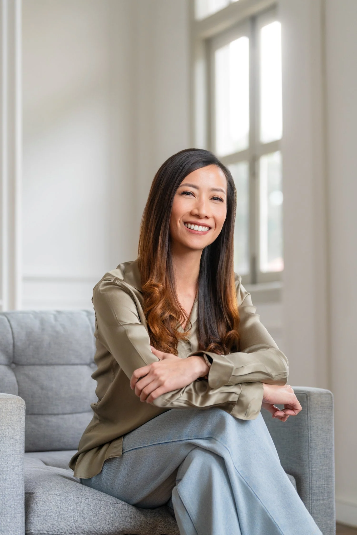 A woman with long dark hair and a warm smile sitting on a gray couch in a well-lit room with large windows, wearing a olive satin blouse and jeans.