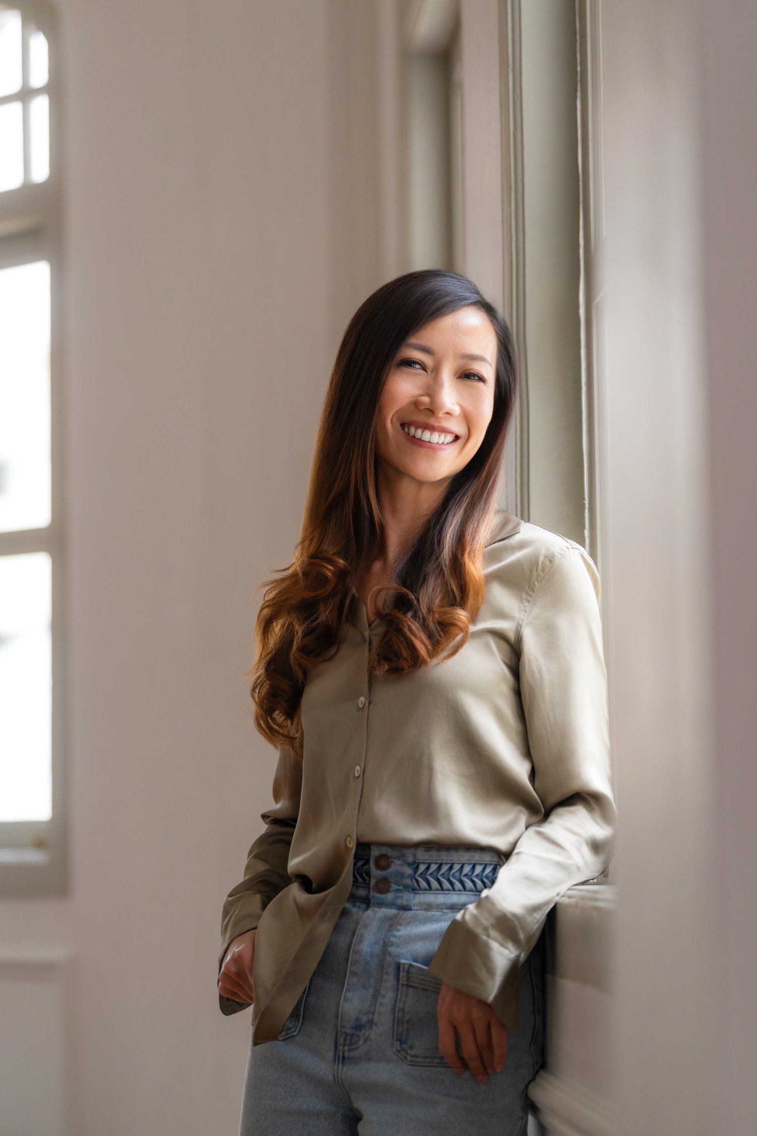 A woman with long brown hair, wearing a beige shirt and jeans, smiling and standing in front of a window.