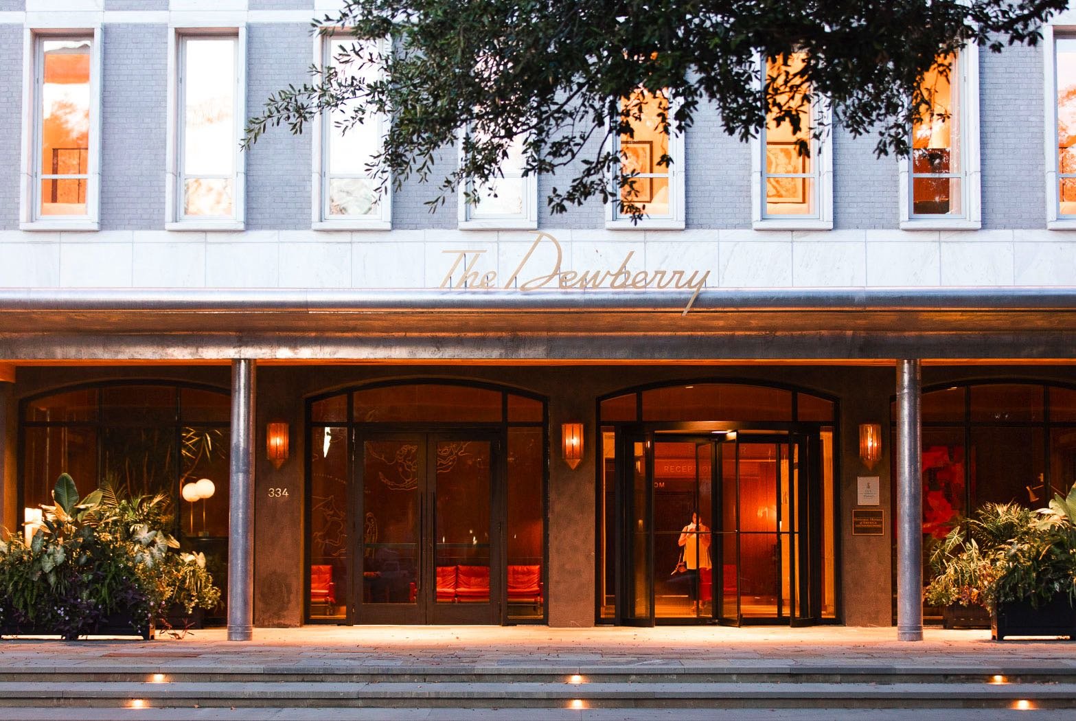 Front entrance of The Dewberry hotel with glass doors, warm lighting, and potted plants on either side, illuminated steps leading to the entrance, and a person visible through the glass.