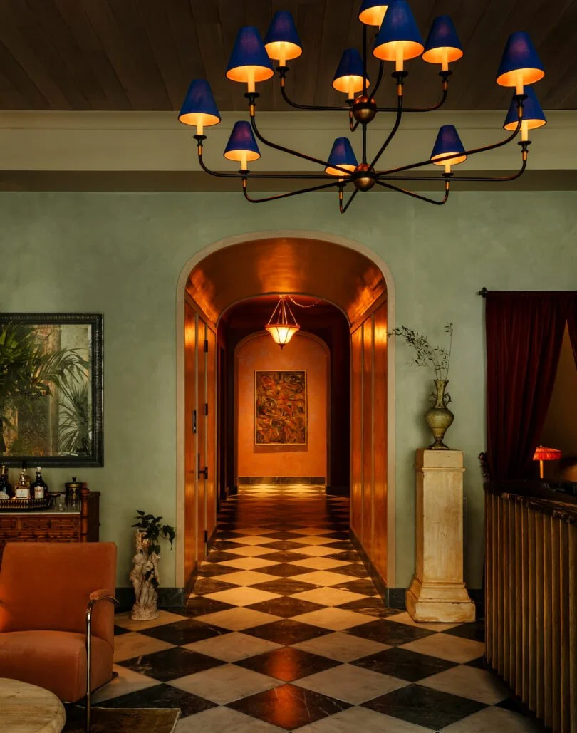 Elegant interior with a checkered black and white marble floor, a chandelier with blue lampshades hanging from a wooden ceiling, and a hallway leading to a warmly lit area with artwork on the wall.