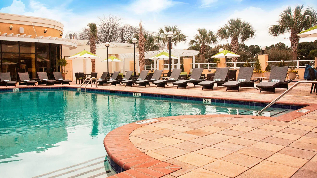 Empty outdoor pool area with lounge chairs, umbrellas, palm trees, and a building in the background during daytime.