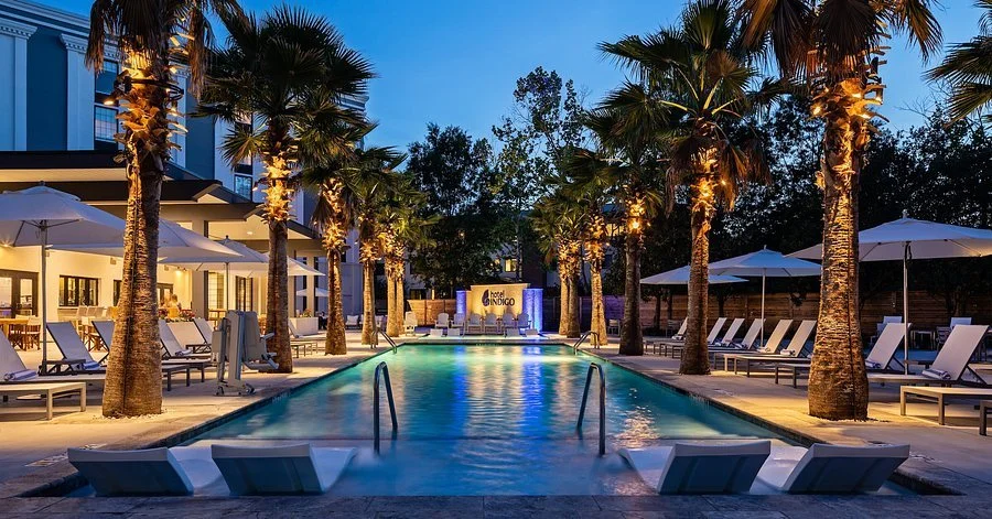 Outdoor hotel swimming pool at dusk, with palm trees, lounge chairs, umbrellas, and lighting