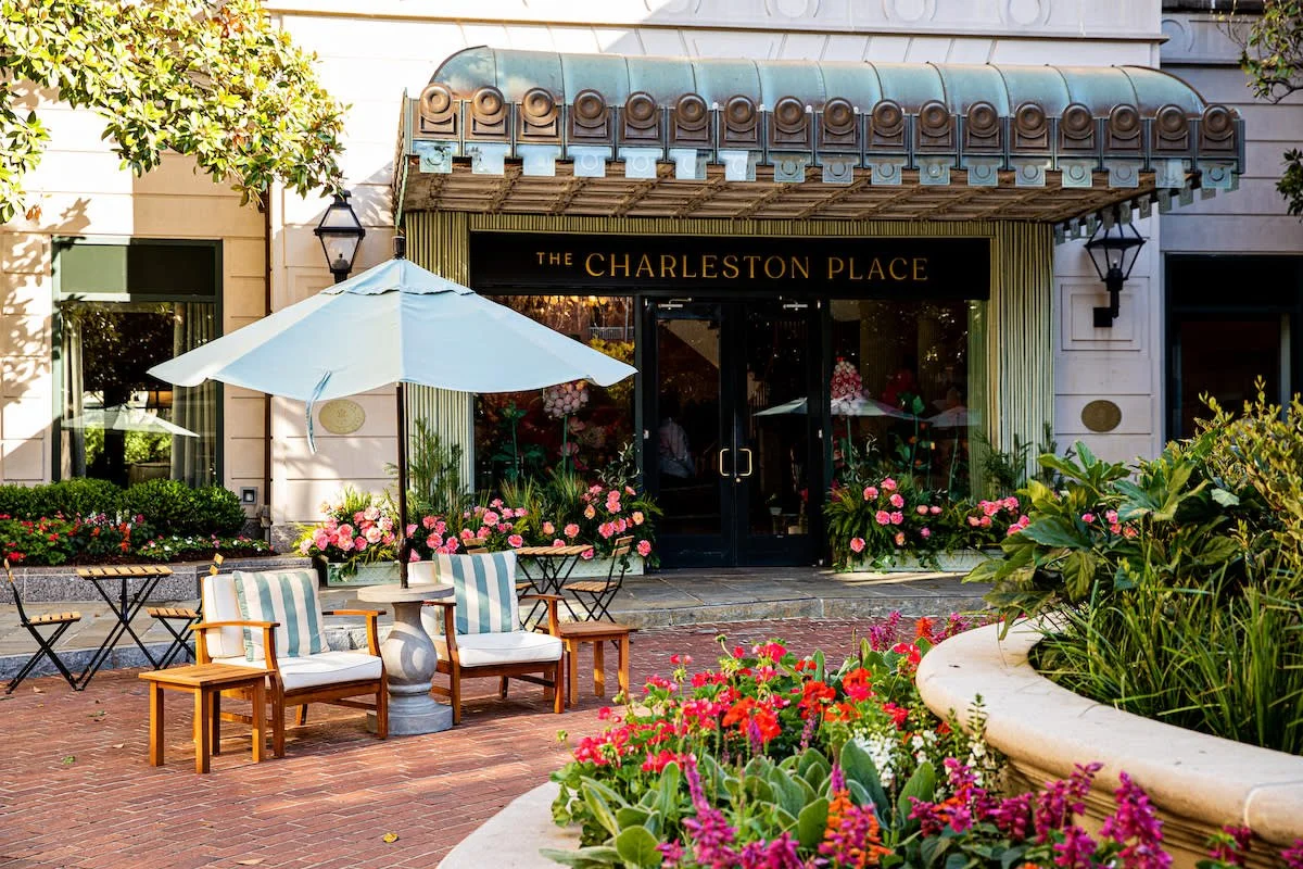 Exterior view of The Charleston Place, a hotel or building, with a decorative awning above the entrance, outdoor seating with chairs, tables, umbrellas, and lush flower beds in front.
