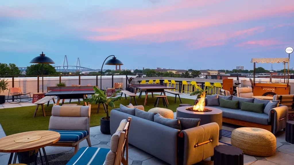 Rooftop outdoor lounge with lounge chairs, sofas, a fire pit, and string lights against a city skyline at sunset.