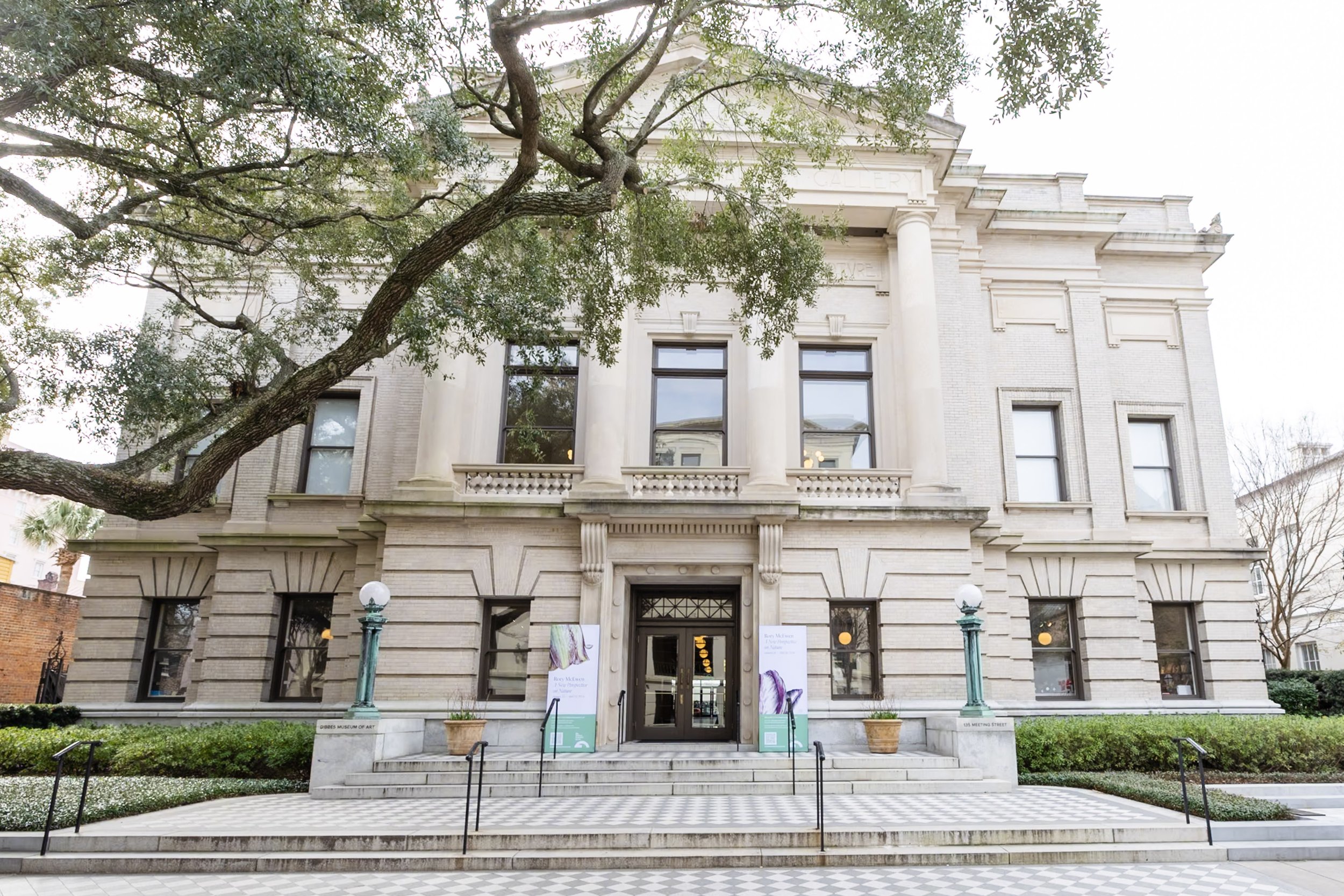 Front view of a historic beige stone museum building with columns, large windows, and steps leading up to the entrance, surrounded by greenery and trees.