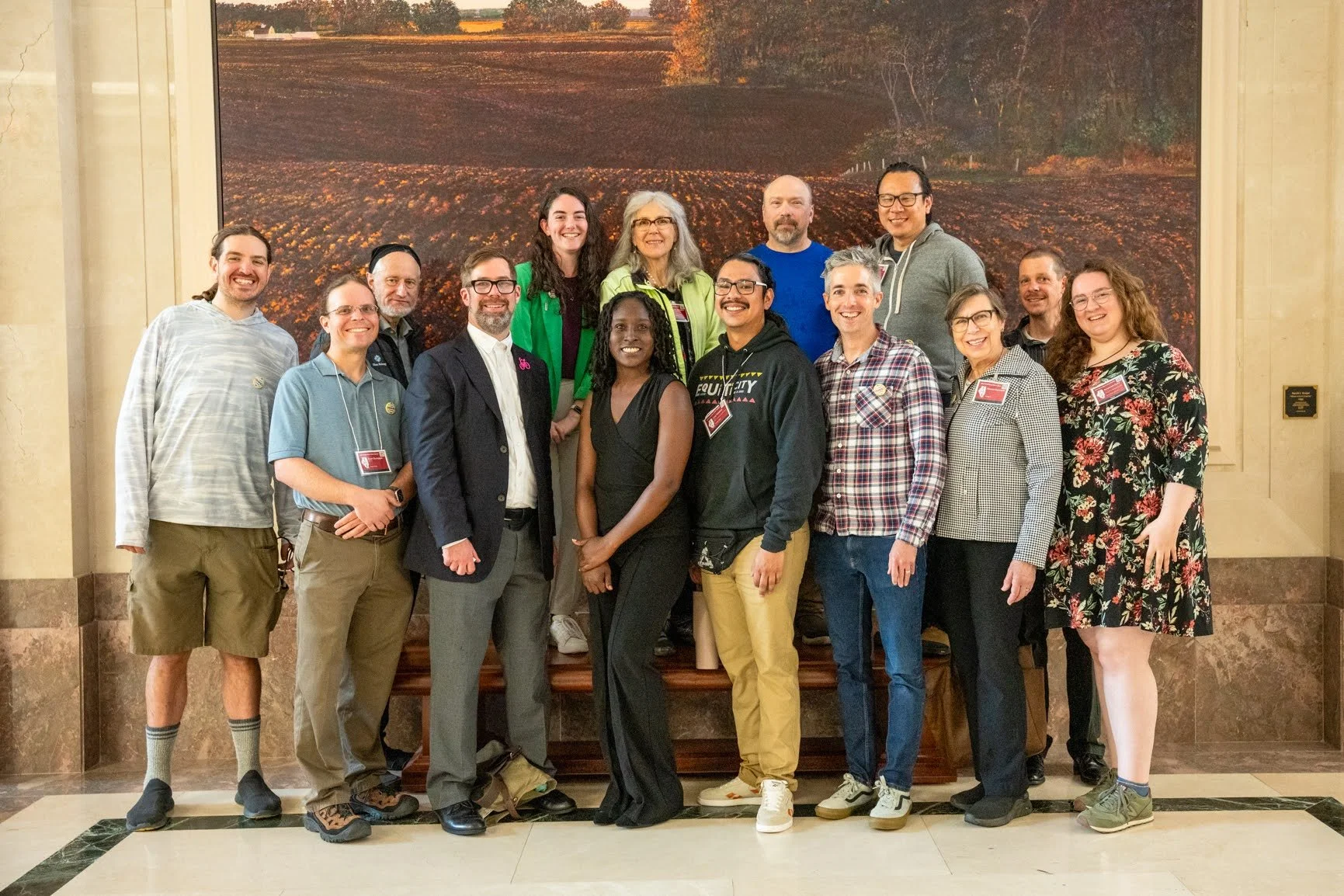 Group of diverse people standing together indoors in front of a large landscape painting, smiling at the camera.