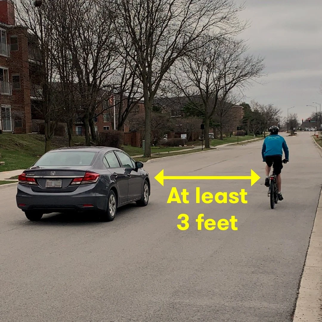 Dark grey sedan pictured next to a bicycle on a side street.  There is bright yellow text along with a line with two arrows on each end stating "at least 3 feet".