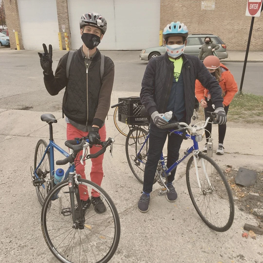Two young men seen wearing masks, and helmets next to their bikes.  Both smiling and the gentleman on the left is making a "peace" sign with right hand.