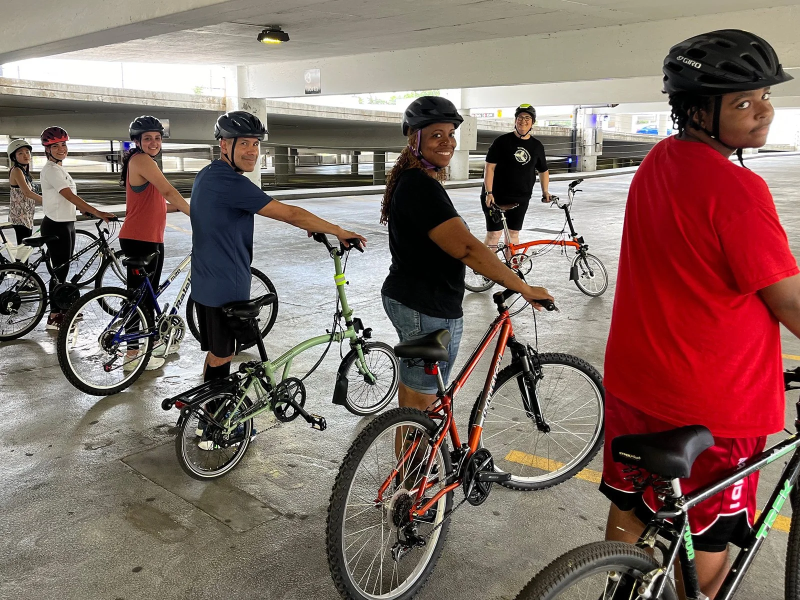 Group of six people, including women and men, wearing helmets and casual clothes, standing with bicycles in an underpass parking garage.