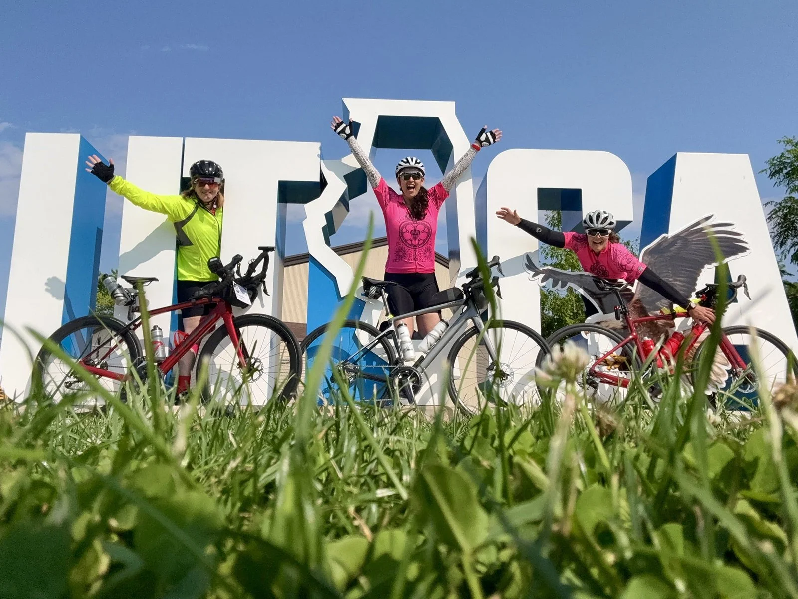 Three women in cycling gear and helmets joyfully posing with their bikes in front of large white letters, with a blue sky and green grass in the background.