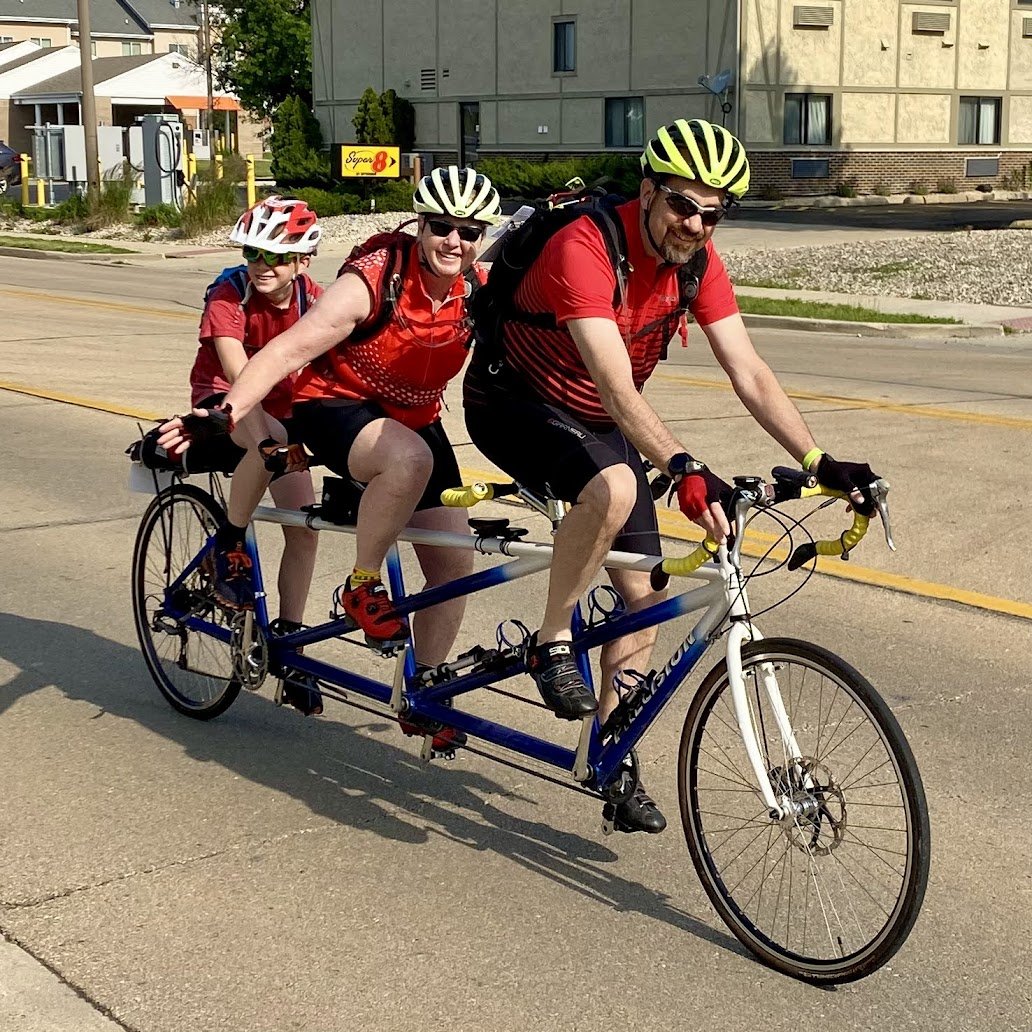 Three individuals pictured smiling on a three person tandem bike smiling at the camera.