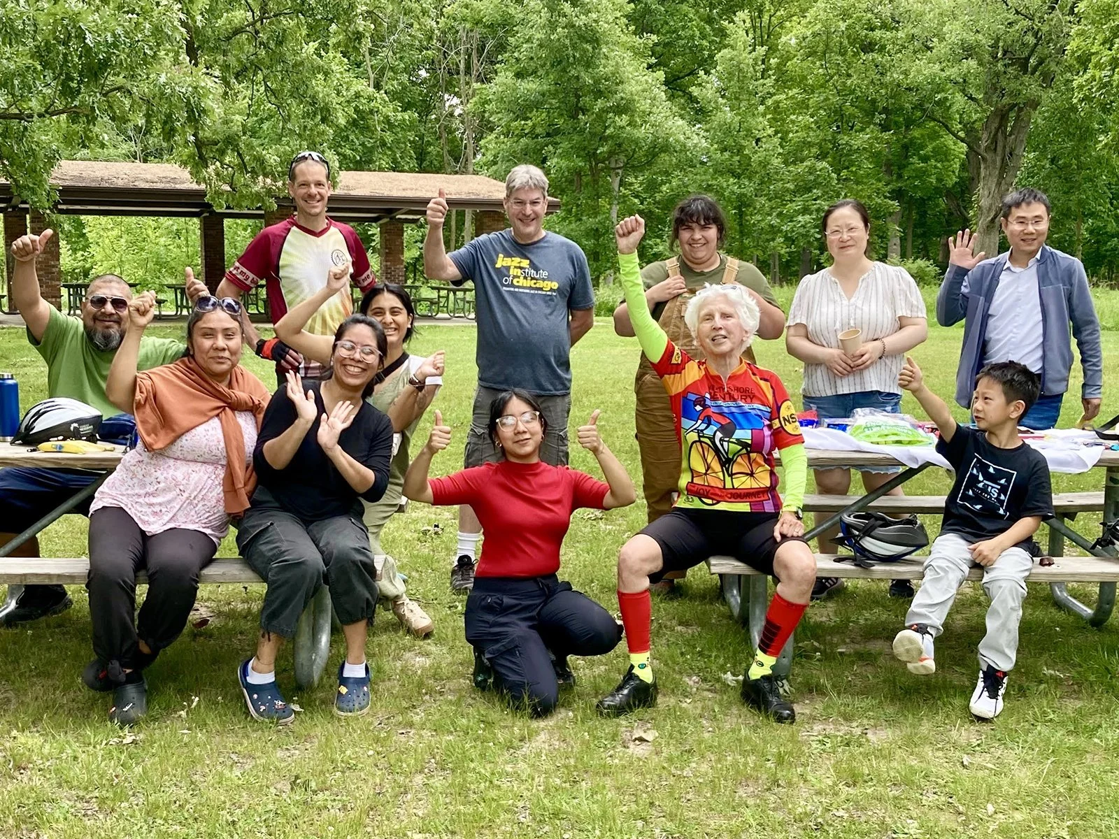 Group of people outdoors in a park, smiling, raising their hands, and cheering. Some are sitting on benches, others standing, with trees and a pavilion in the background.