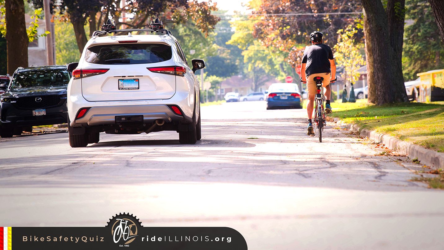 A person riding a bicycle on a residential street with parked cars and trees lining the sidewalk.