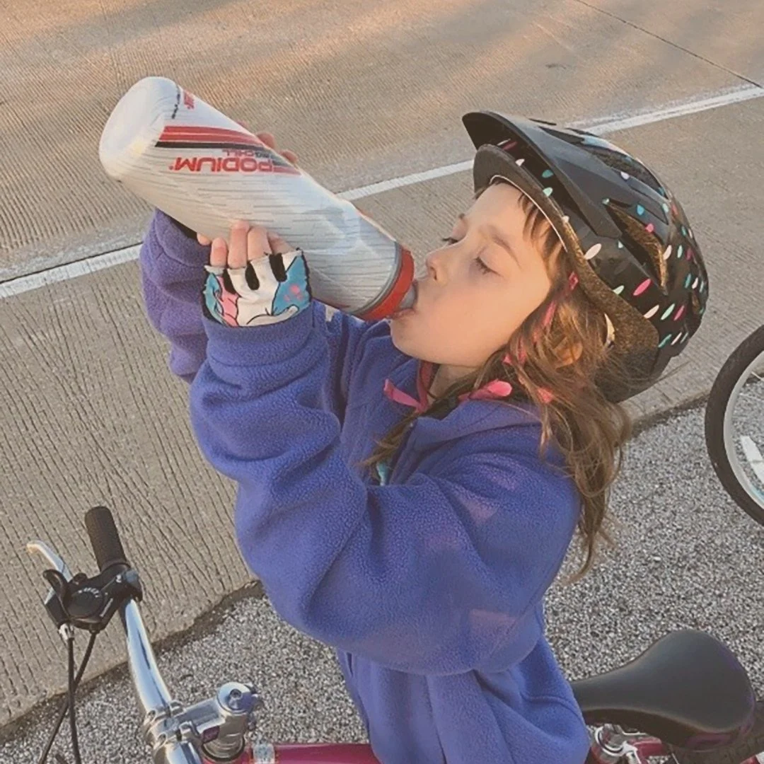 Young girl pictured with a helmet drinking out of a sports water bottle.  She can be seen with her red bike.