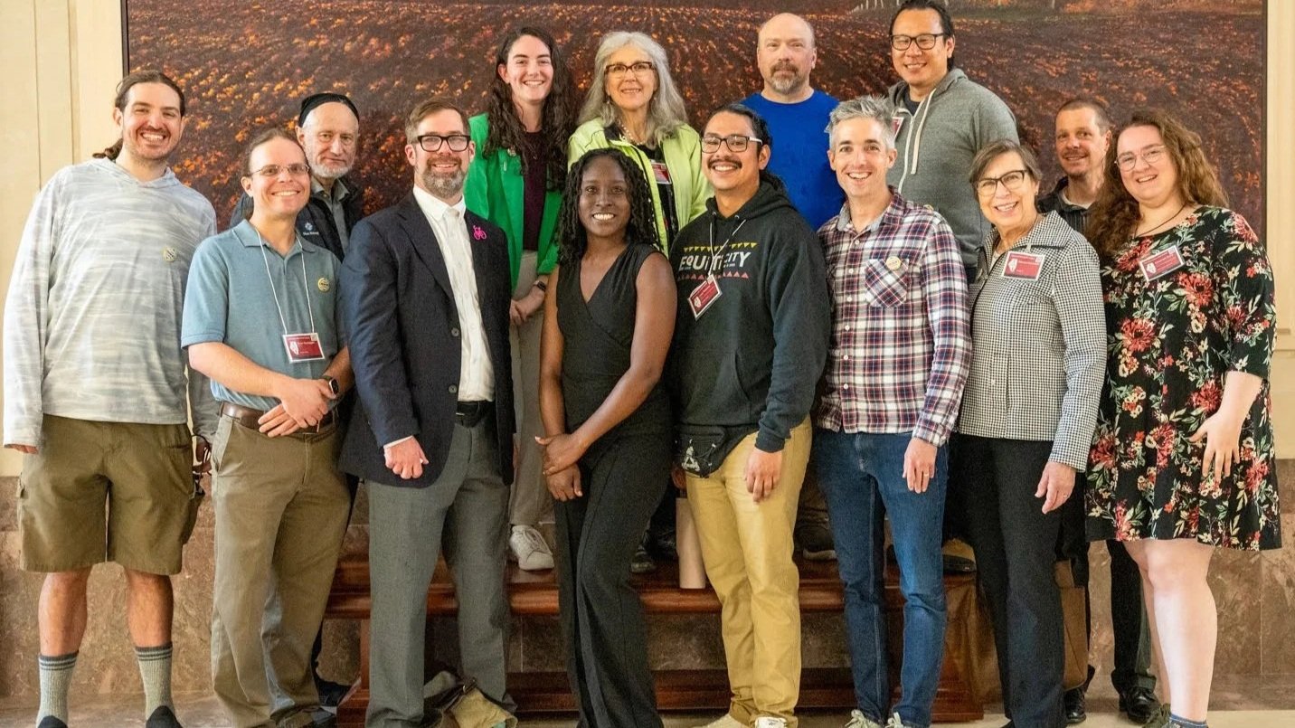 A group of 14 diverse people posing indoors in front of a large framed artwork, smiling for the camera.