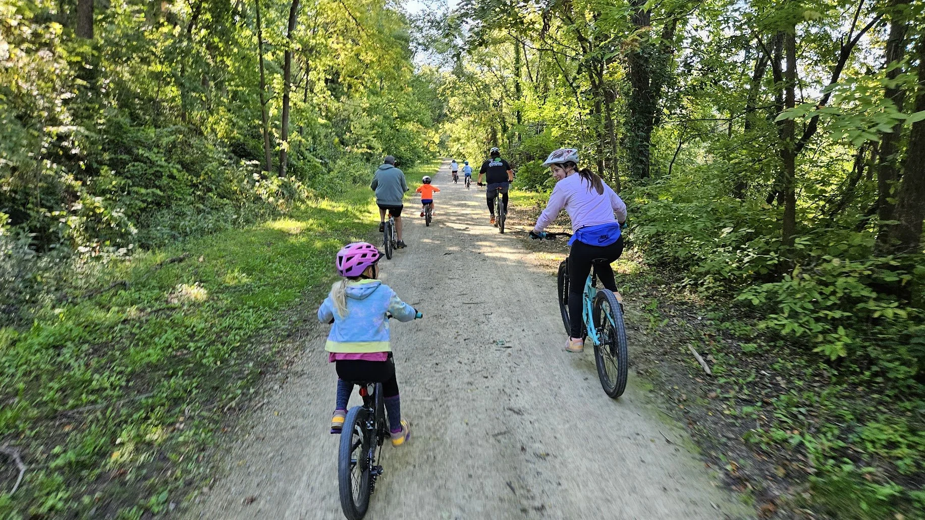 Group of people, including children, riding bikes on a dirt trail through a green wooded area on a sunny day.