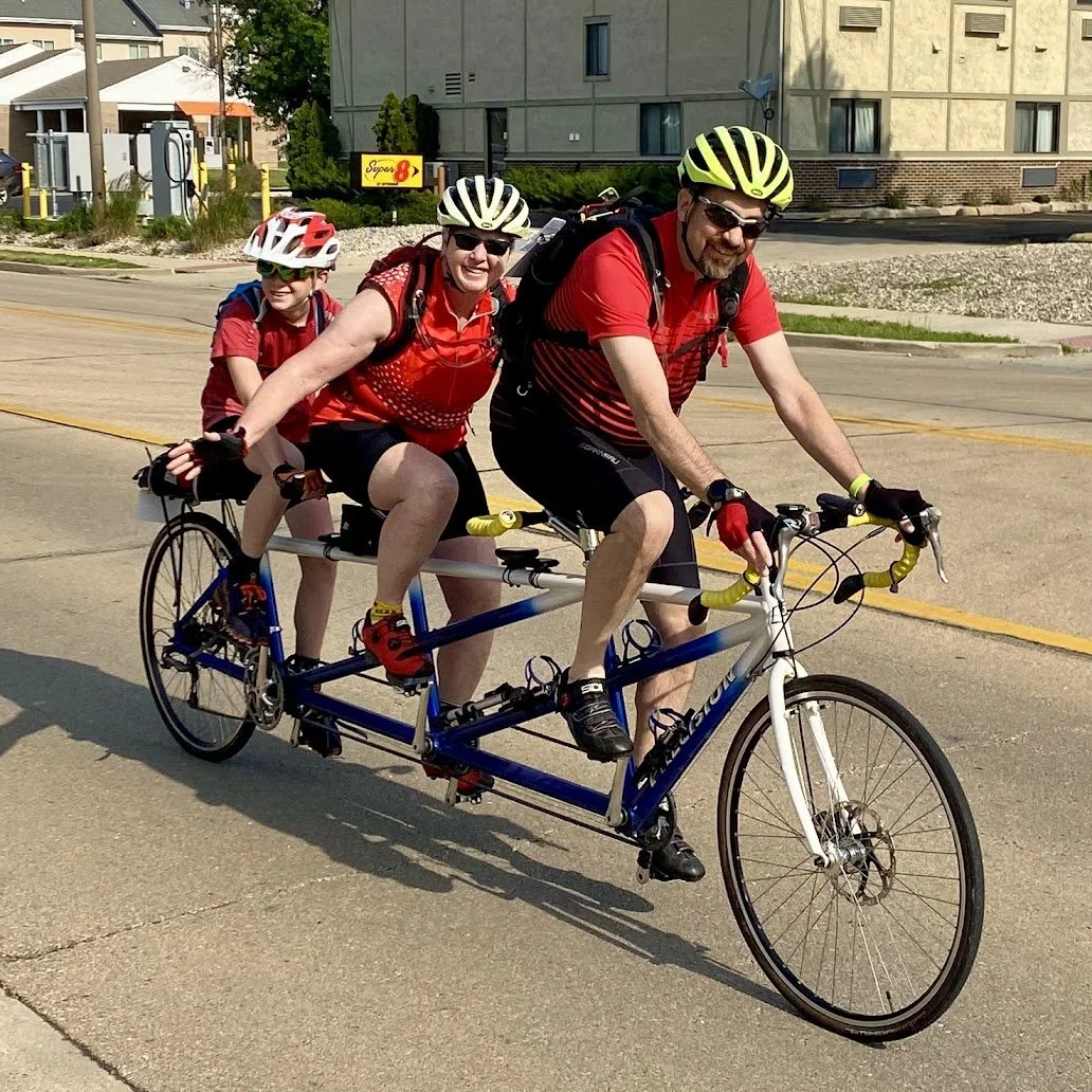 Family in Illinois riding a triple tandem, center rider waving bicycle, they're all wearing matching red jerseys and bike helmets