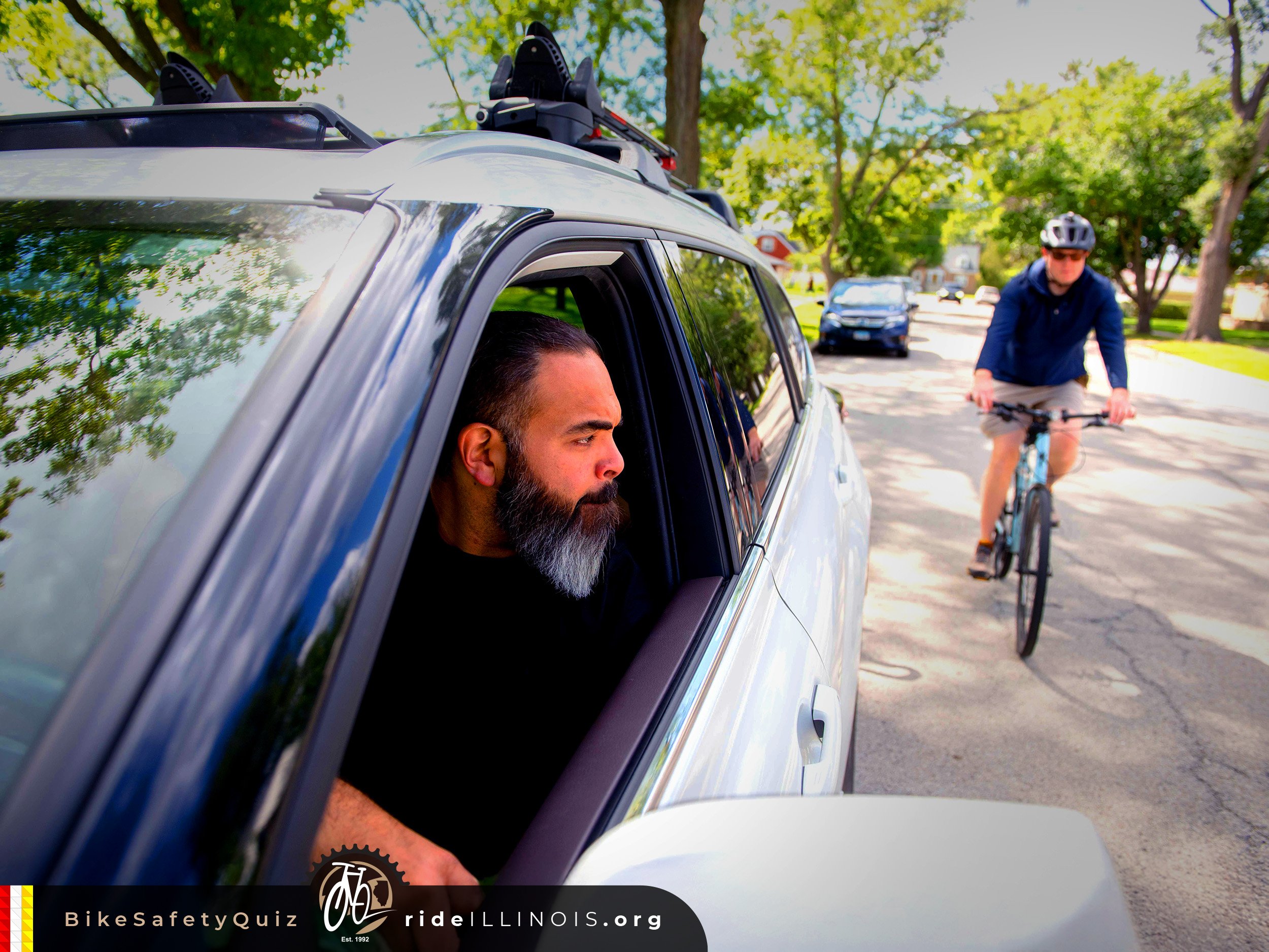 Picture taken up close on the front of the driver's side of a vehicle.  A man is seen looking out the open window over his shoulder.  A bicyclist is seen approaching the vehicle from behind.