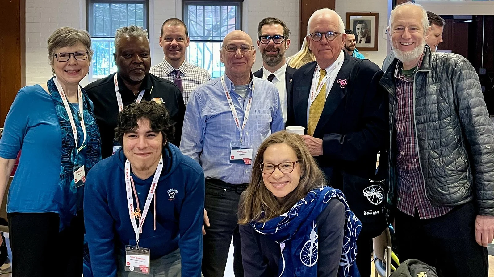 A group of ten diverse people posing for a photo at a conference or event indoors. They are smiling and wearing name badges.