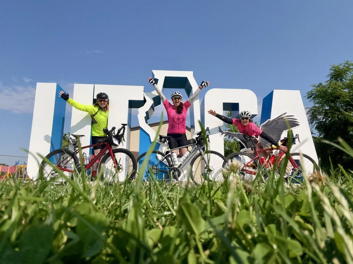 Three woman pictured in bike gear with their bikes popping out of large white statues of different letters.