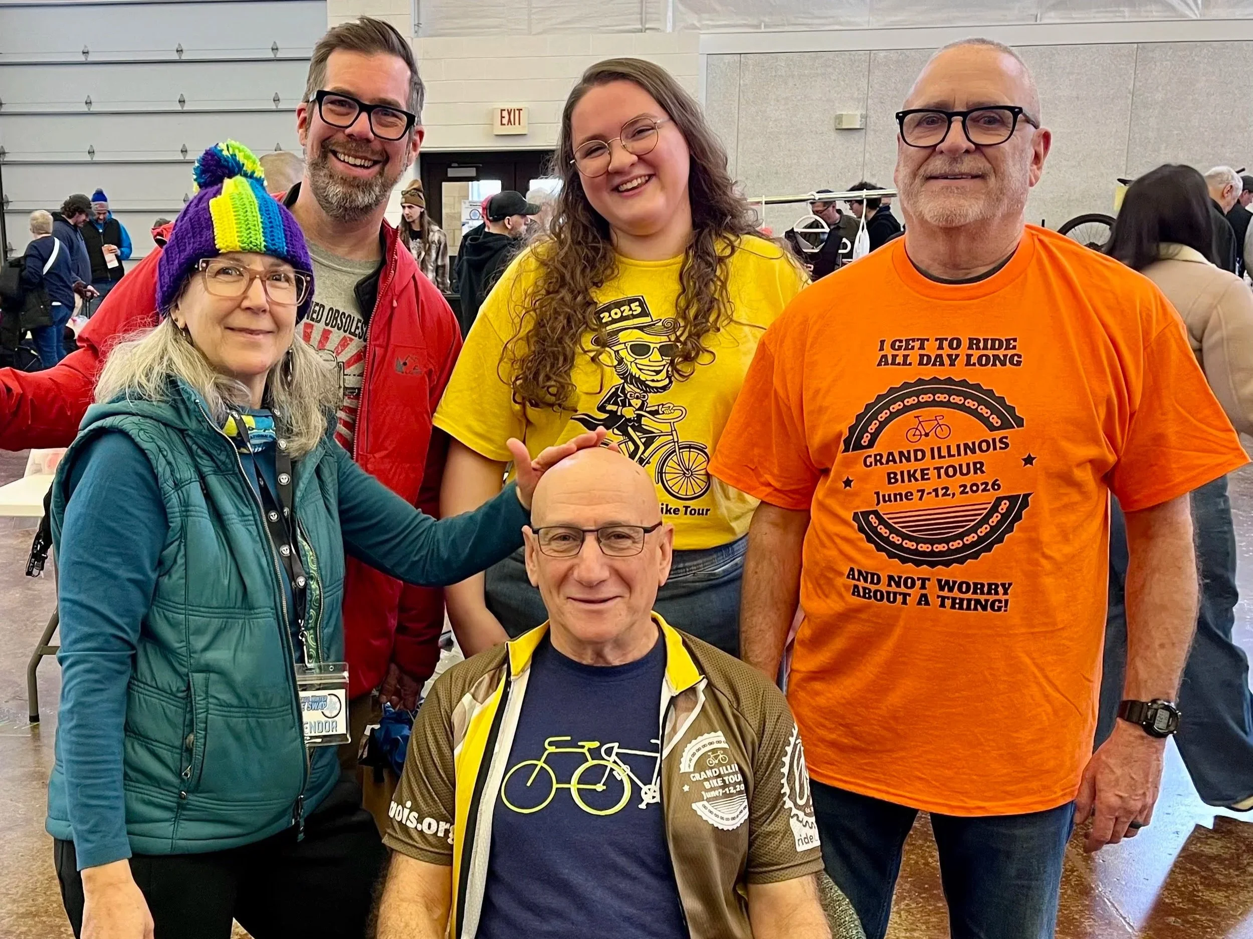 Group of five people smiling at a cycling event, with some wearing cycling-themed shirts and badges, in a large indoor venue.