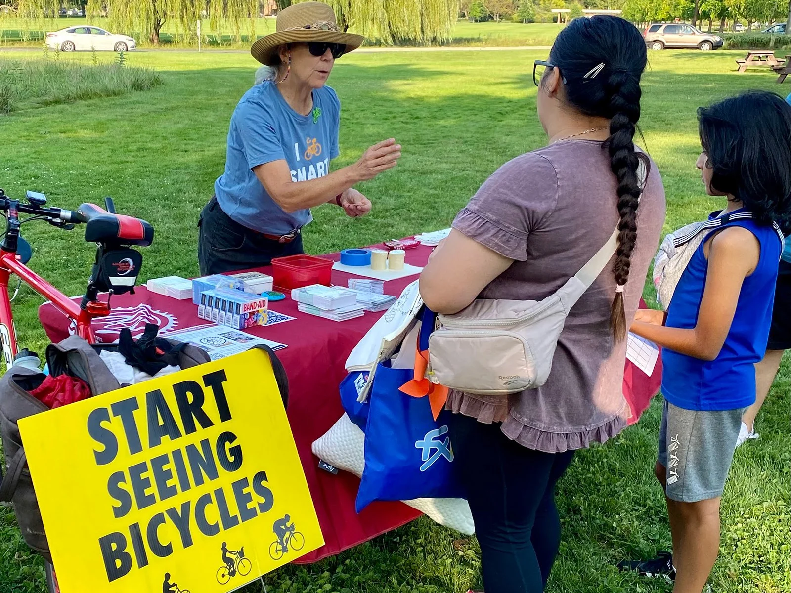 A woman wearing a large sunhat, sunglasses, and a blue t-shirt demonstrating bicycle safety to two girls at an outdoor event. There is a yellow sign that reads 'START SEEING BICYCLES' and a table with brochures, band-aids, and supplies. Green grass and trees are visible in the background.