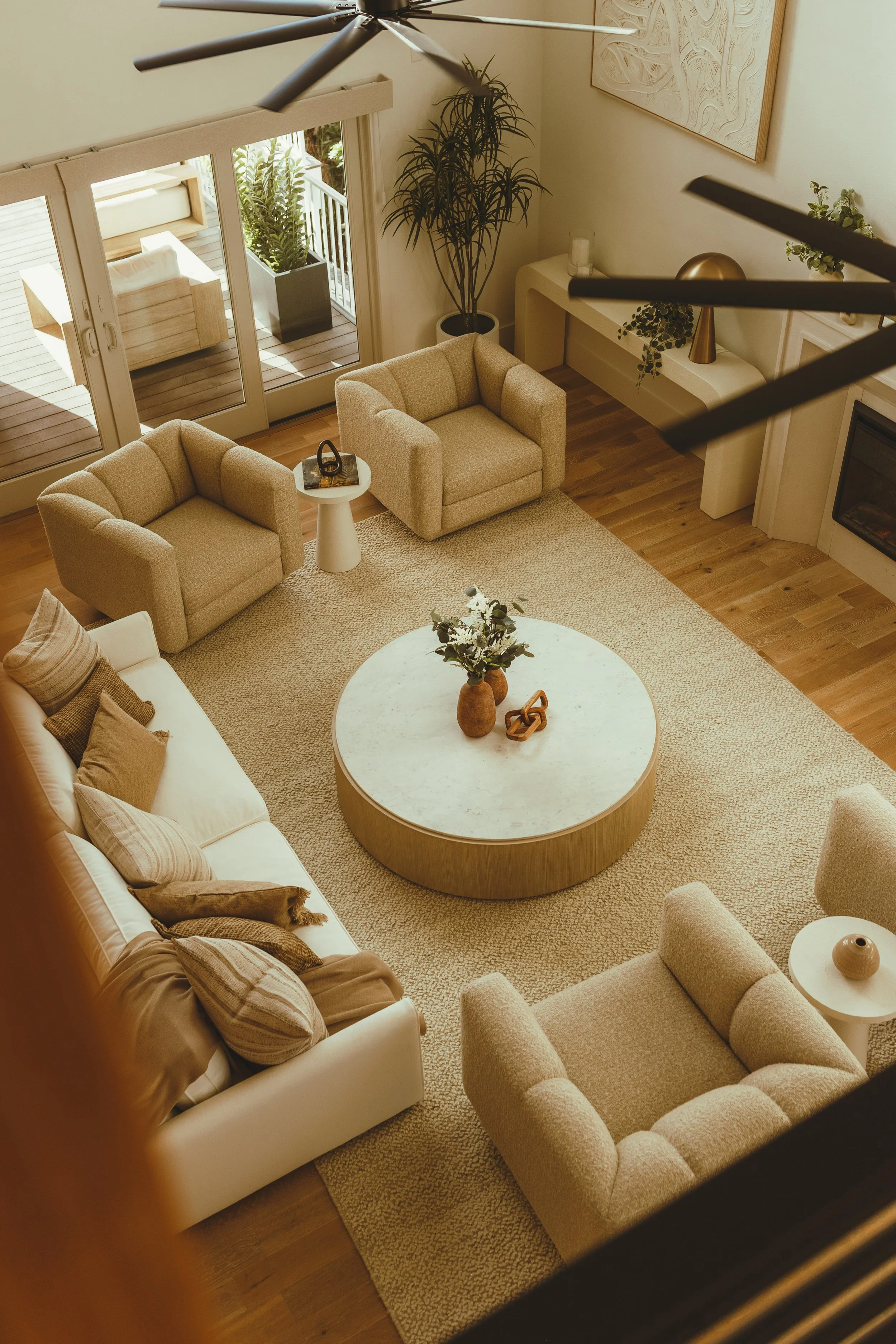 A cozy living room with beige and cream furniture, a round marble coffee table with vases and greenery, a white sofa with pillows, two beige armchairs, large sliding glass doors leading to a balcony with outdoor furniture, and decorative elements like a large plant, artwork, and a golden lamp.