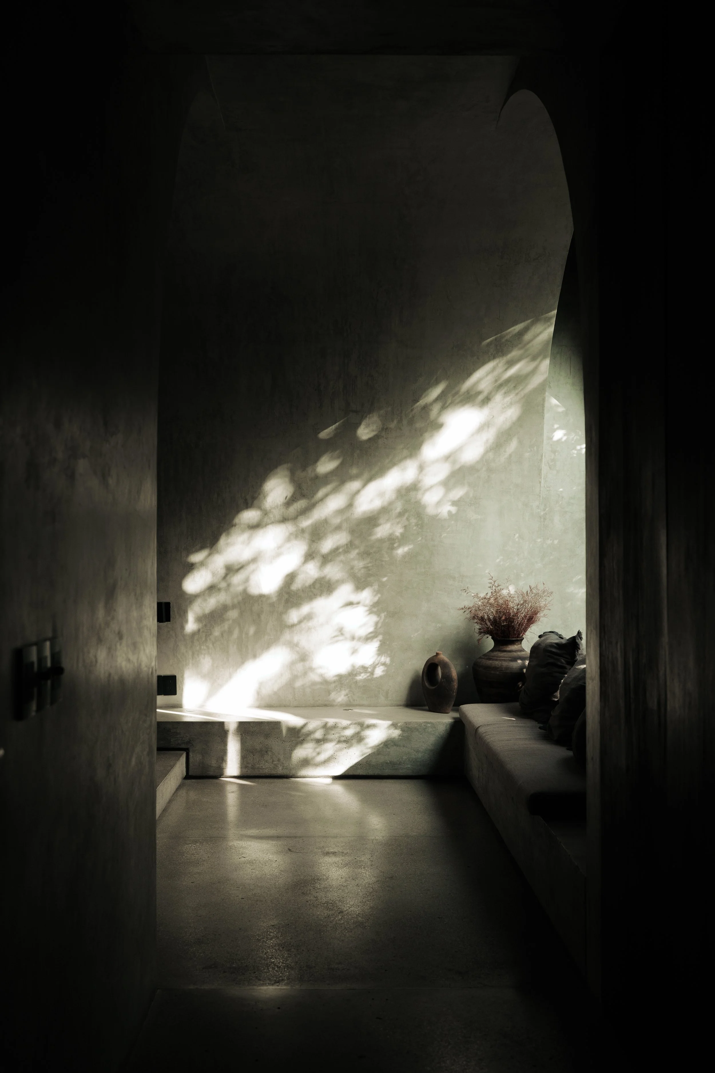 A dimly lit room with sunlight streaming through a window, creating shadows on the wall. The room features a cushioned bench with dark pillows, and large ceramic vases with dried flowers on a ledge.