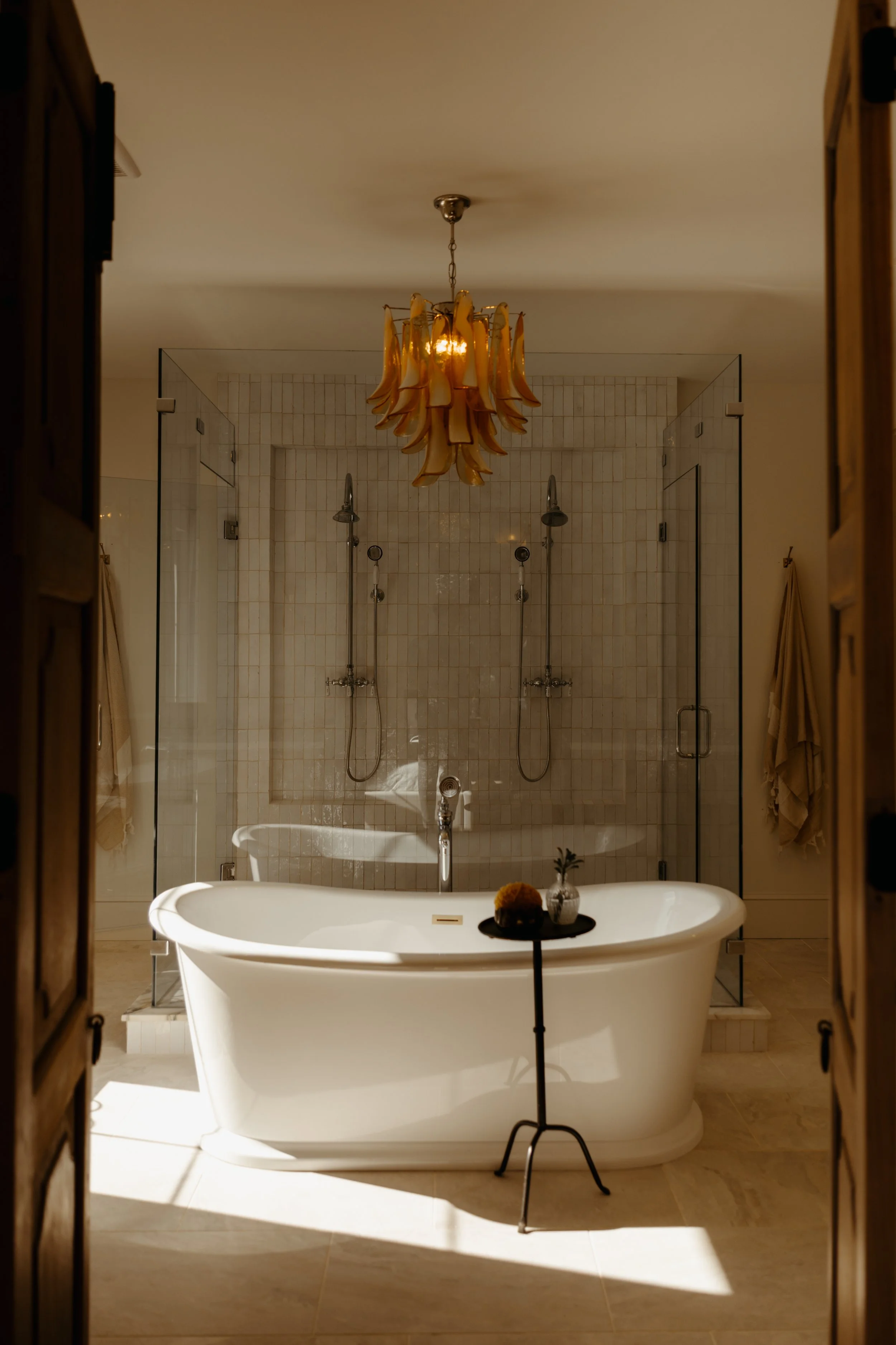 A luxurious bathroom featuring a standalone bathtub with a small black side table holding a small plant and decorative object, behind a glass-enclosed shower with dual shower heads, a vintage chandelier hanging from the ceiling, and warm-toned lighting.