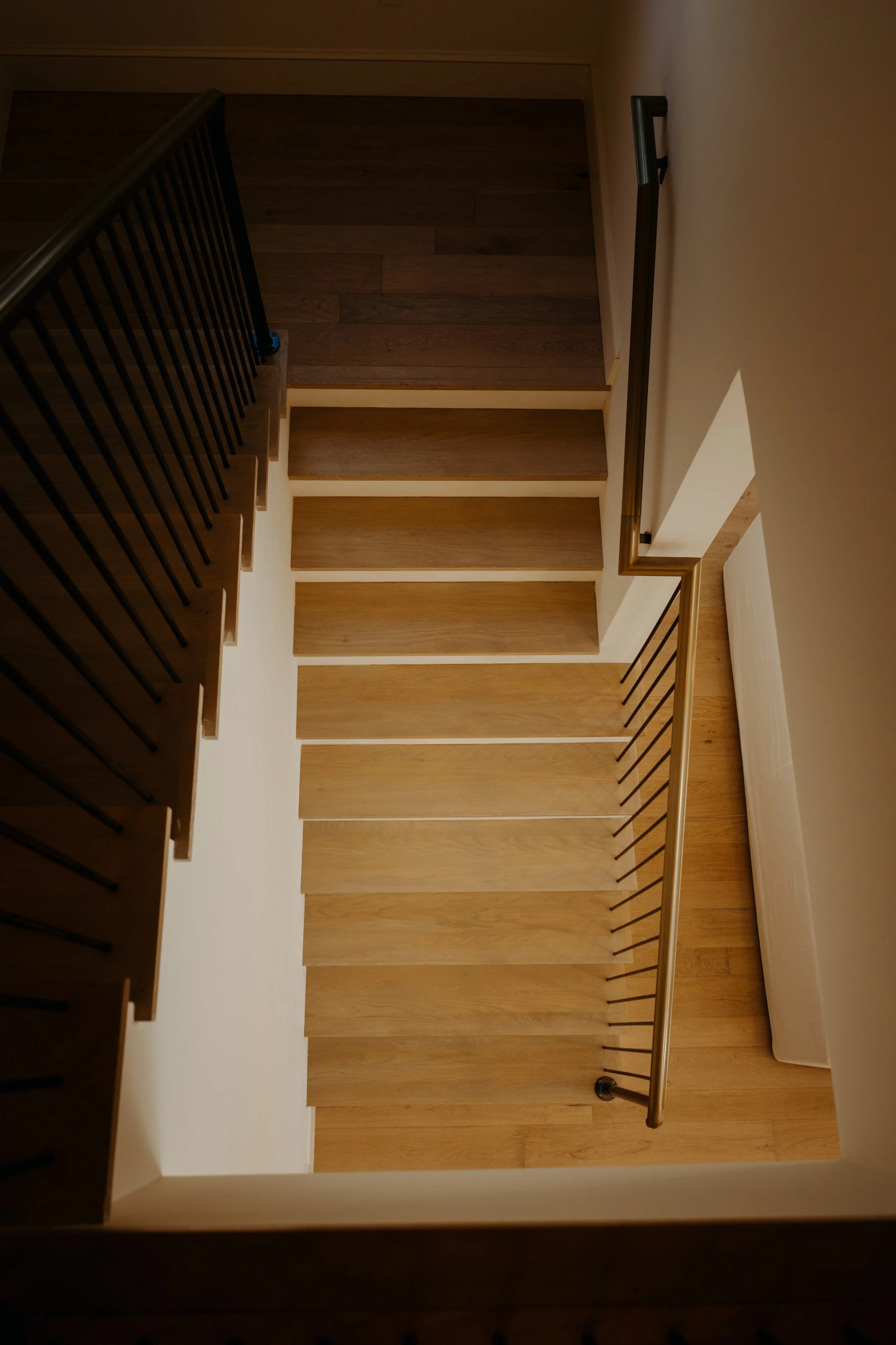 Overhead view of a wooden staircase with black handrails, showing steps with a natural wood finish, ascending from a light-colored wall, with a small section of dark flooring visible at the top.