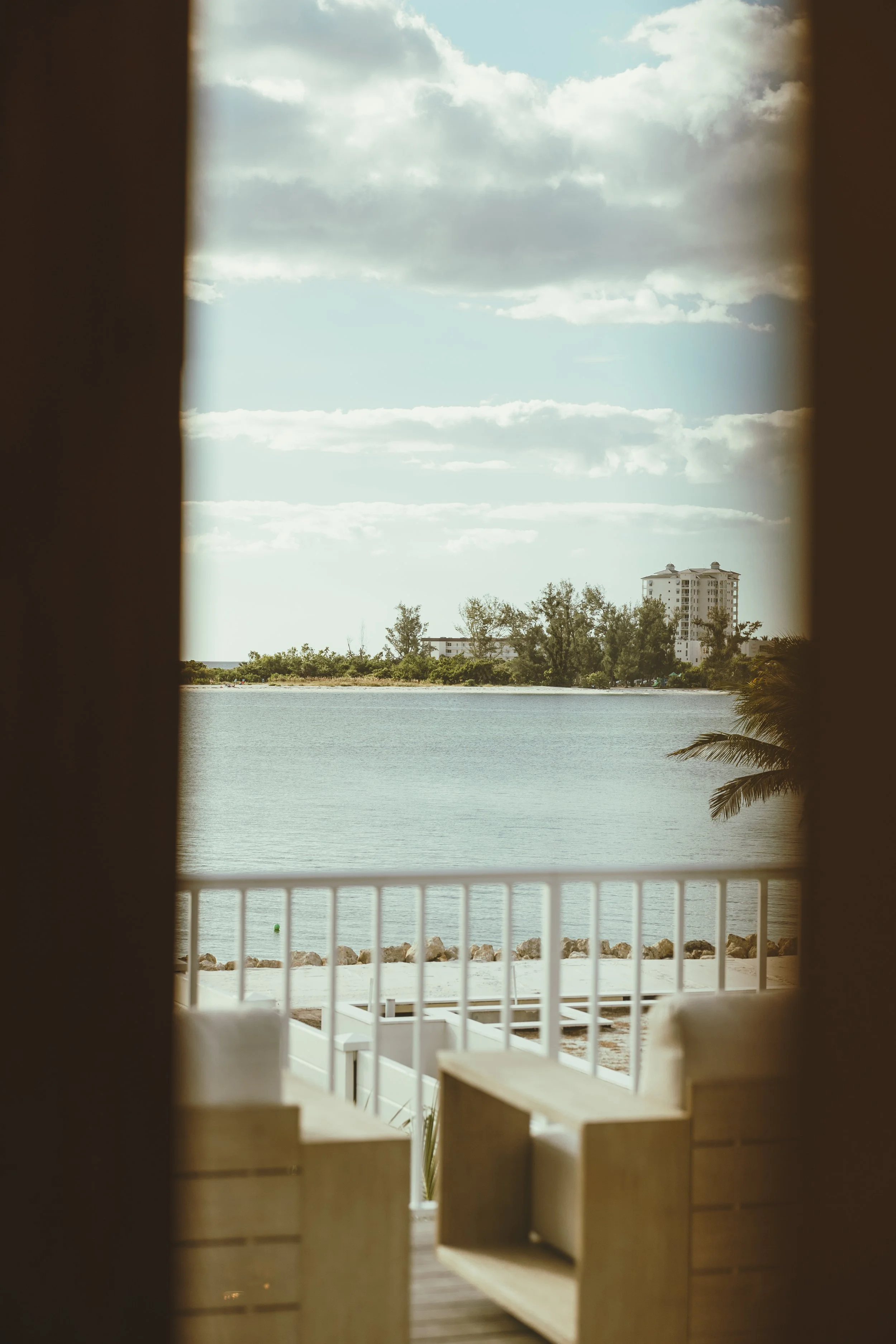 View of a calm body of water seen from a window or doorway, with a sandy shoreline and rocks in the foreground, and trees with a building in the distance under a partly cloudy sky.