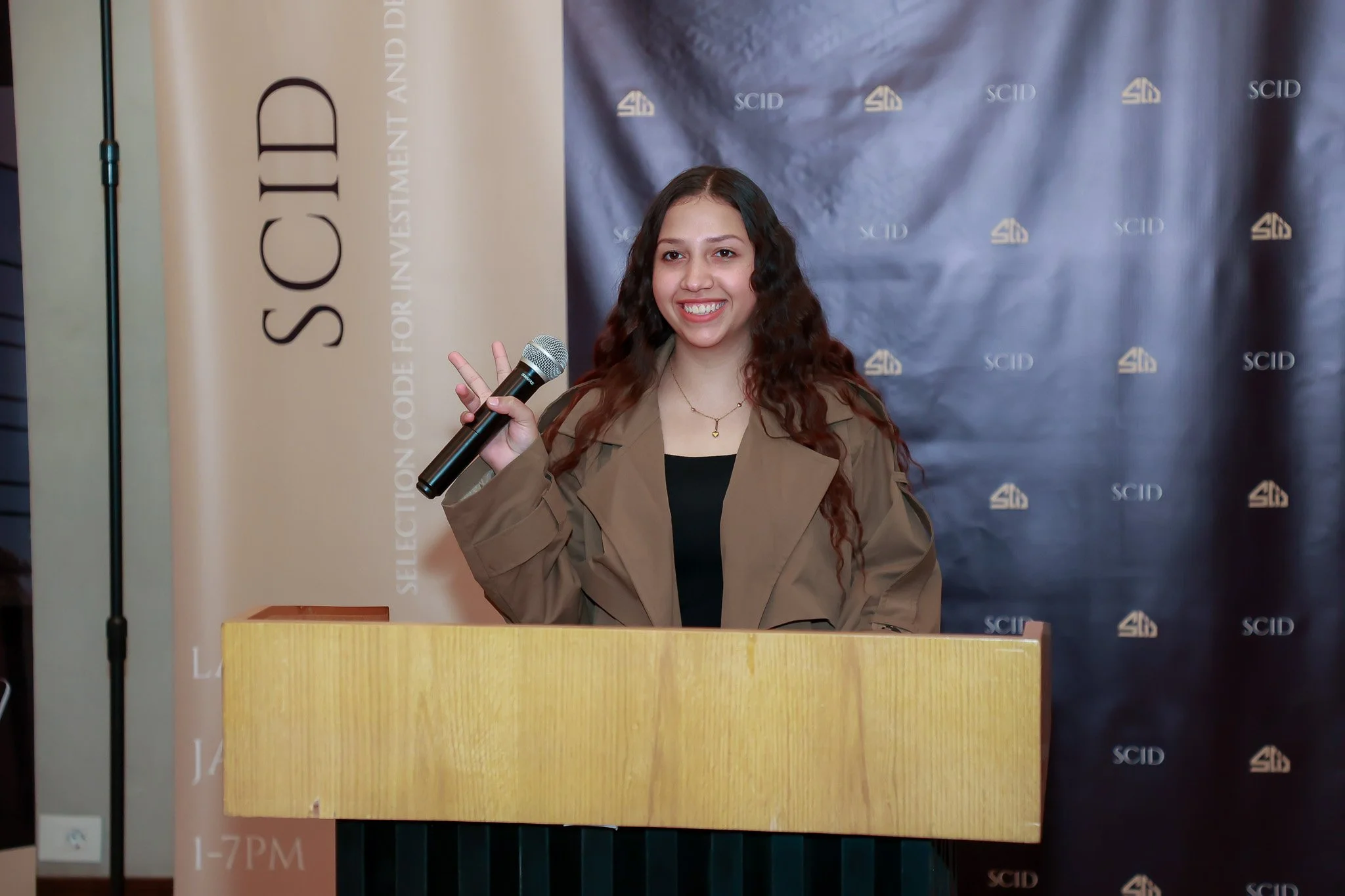 A young woman with long curly hair smiling, holding a microphone and making a peace sign with her right hand, standing behind a wooden podium at an indoor event.
