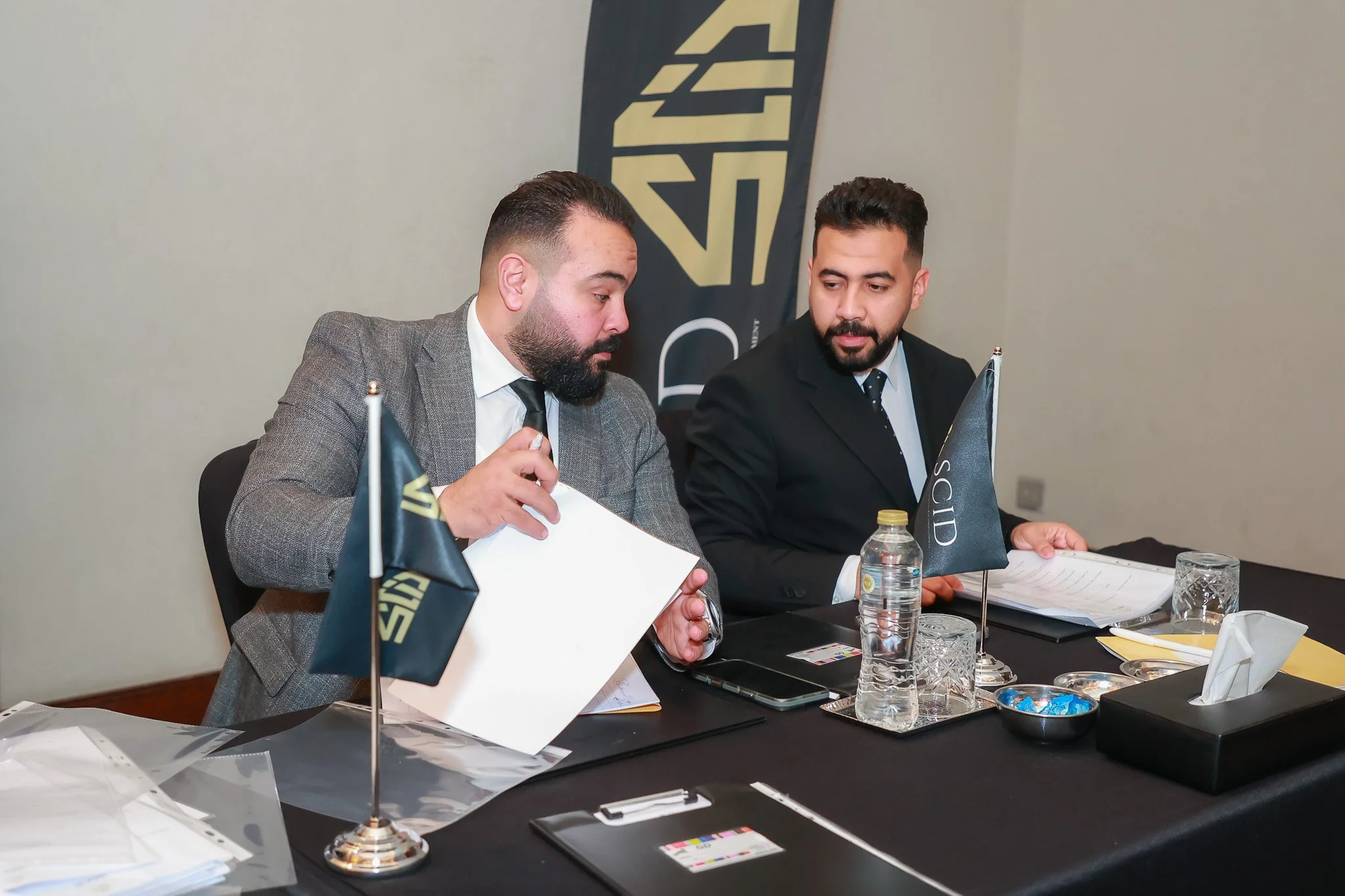 Two men in suits sitting at a conference table with flags, papers, a water bottle, glasses, and a tissue box, engaged in discussion during a business meeting.