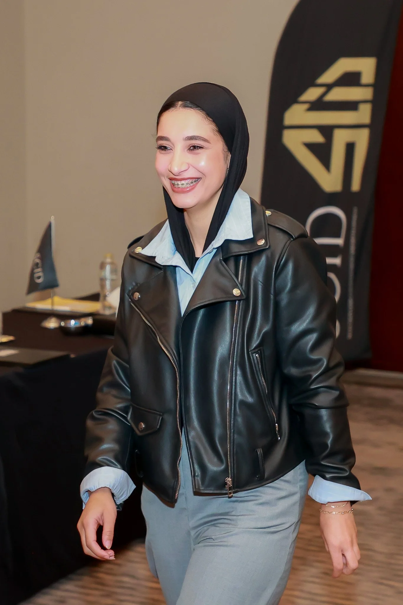Young woman smiling, wearing a black leather jacket over a blue collared shirt, with a black headscarf and light-colored pants, indoors at a conference or event.