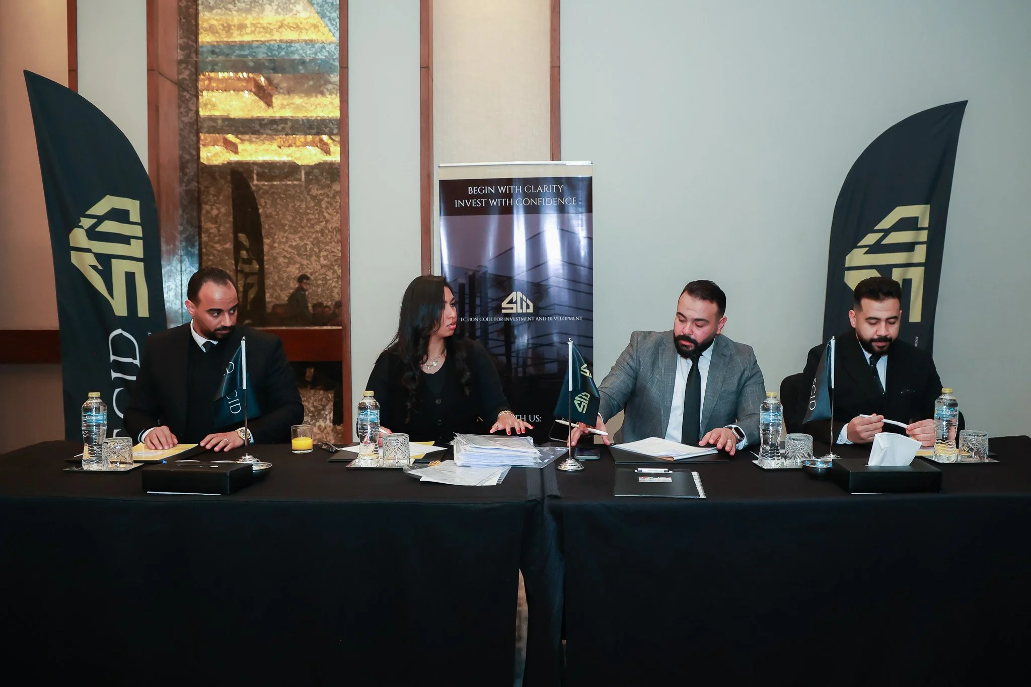 Four business professionals, three men and one woman, sitting at a black conference table with documents, bottles of water, and glasses, in a corporate meeting room. Two black flags with a white logo are on either side, and a banner behind them displ