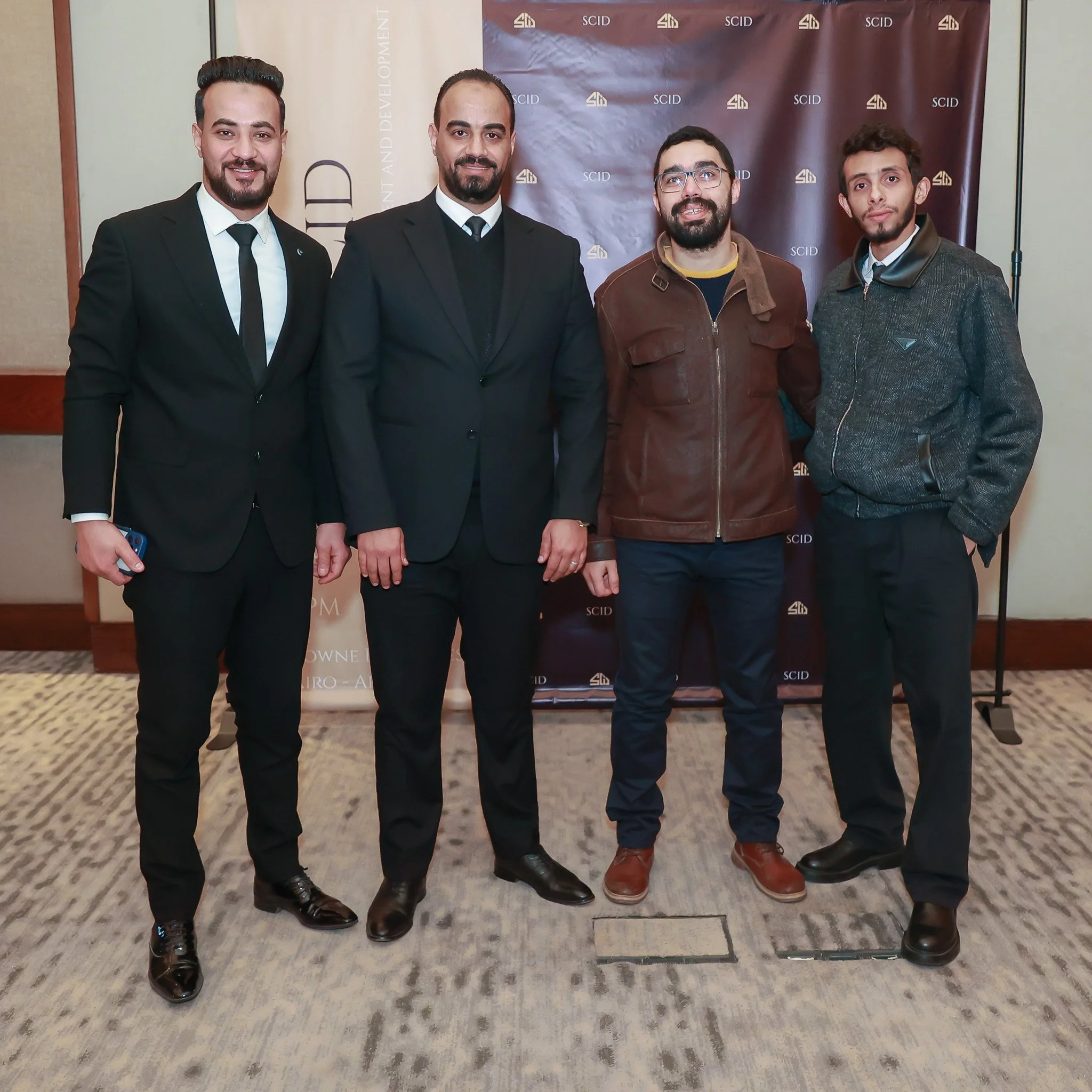 Four men standing in front of a backdrop with logos at an event. Three are dressed in suits, and one in casual attire with a leather jacket.