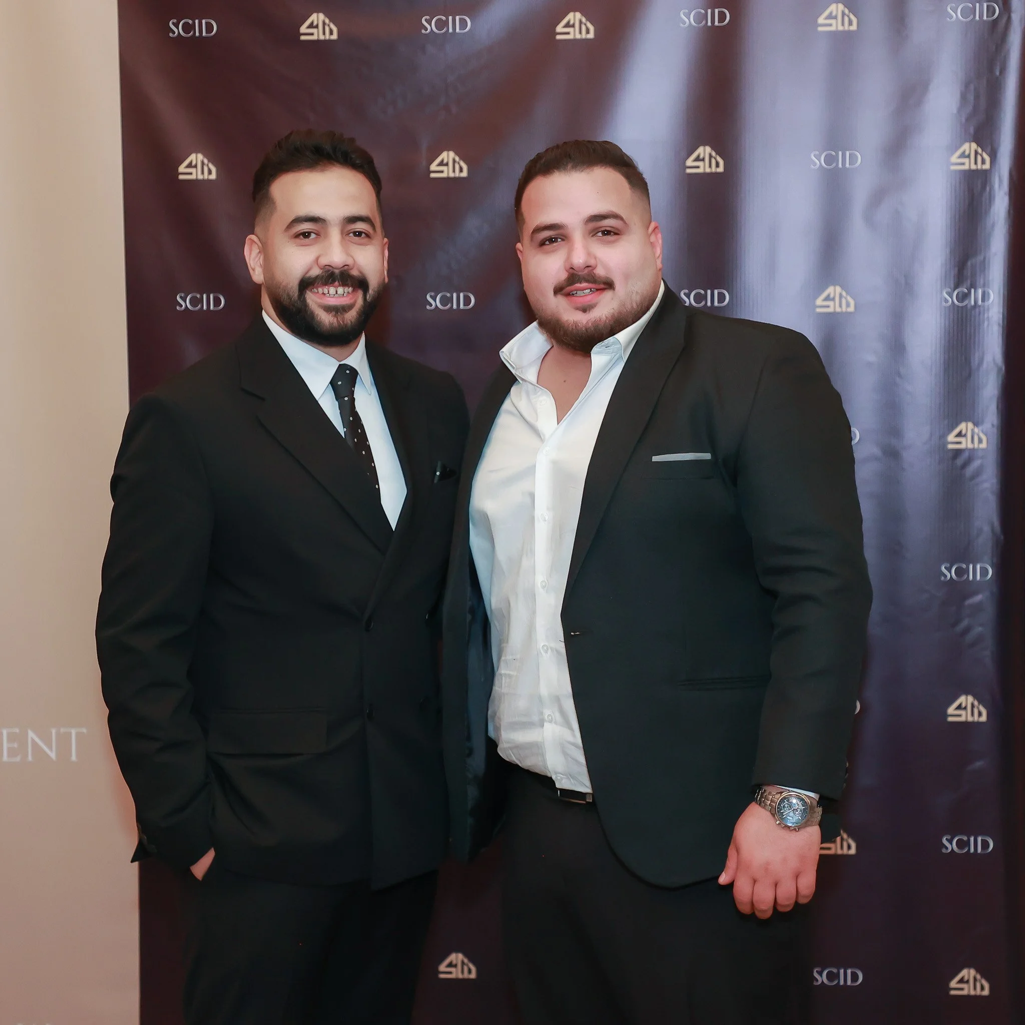 Two men dressed in black suits and white shirts standing in front of a backdrop with 'SCID' logos, smiling for the photo.