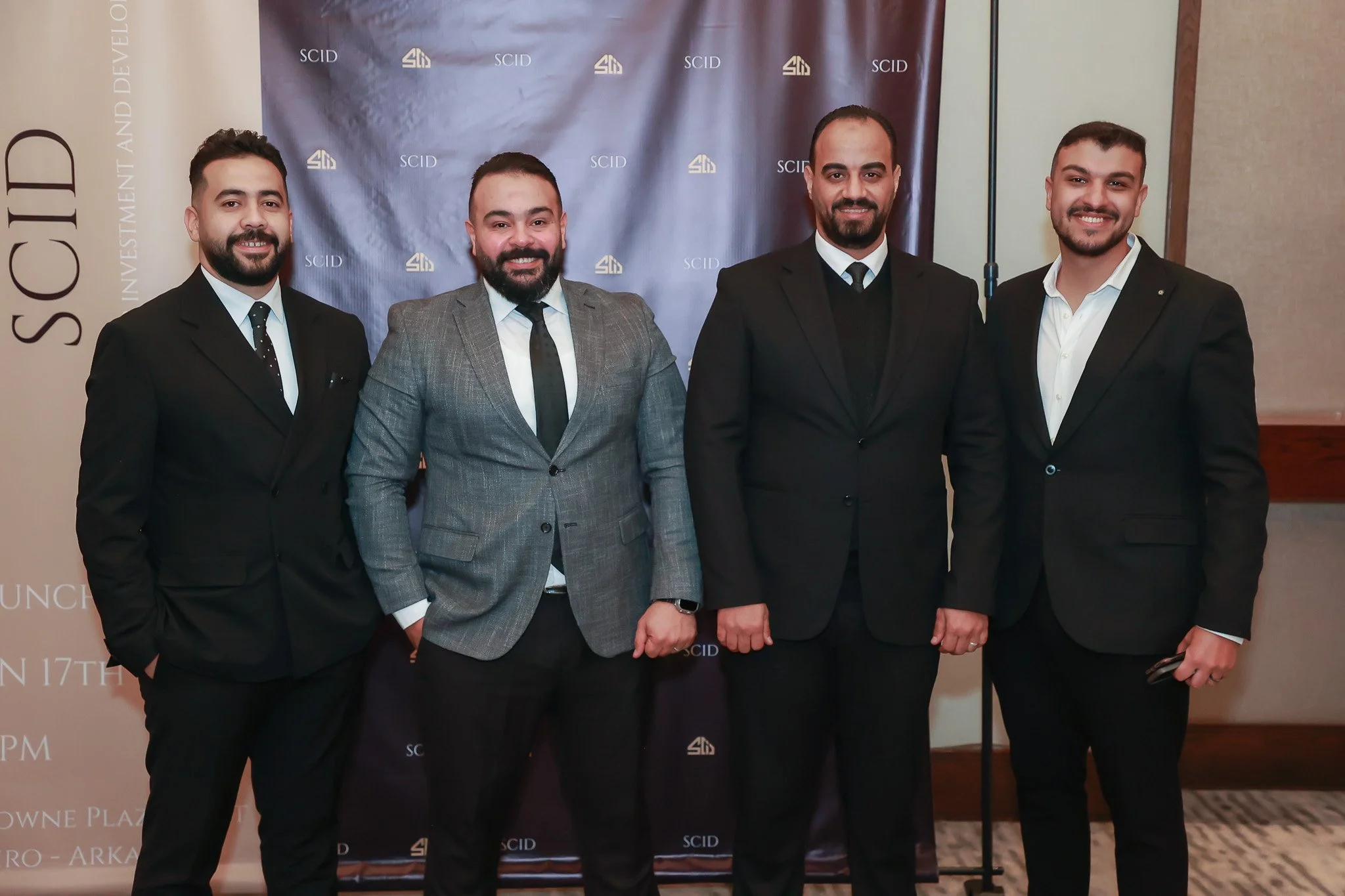 Four men in suits standing together indoors, posing for a photo at a formal event, with a backdrop displaying the logos 'SCID' and a conference or event information banner.