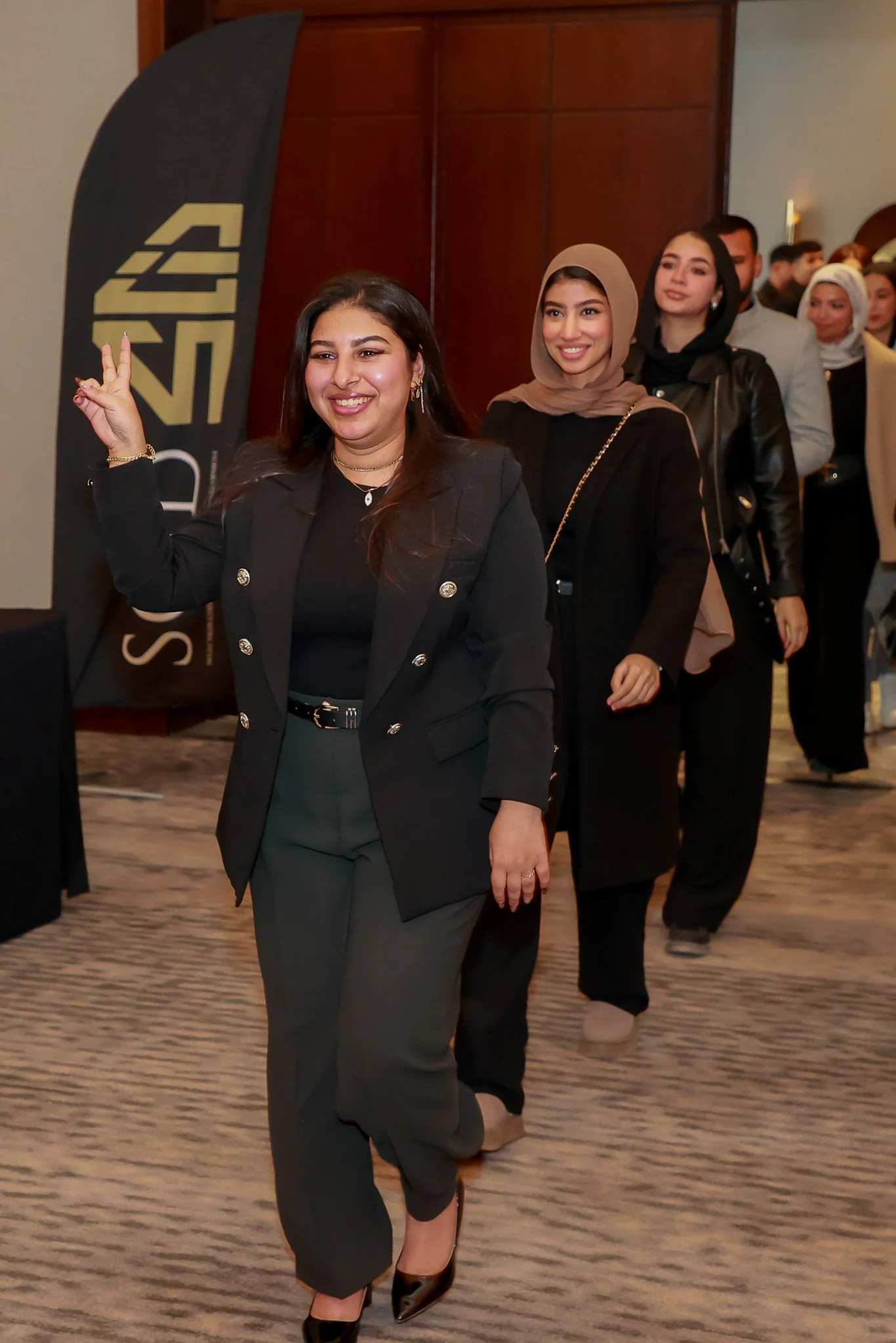 Group of diverse women, dressed in formal attire, walking in a line at an indoor event, with one woman smiling and making a peace sign.
