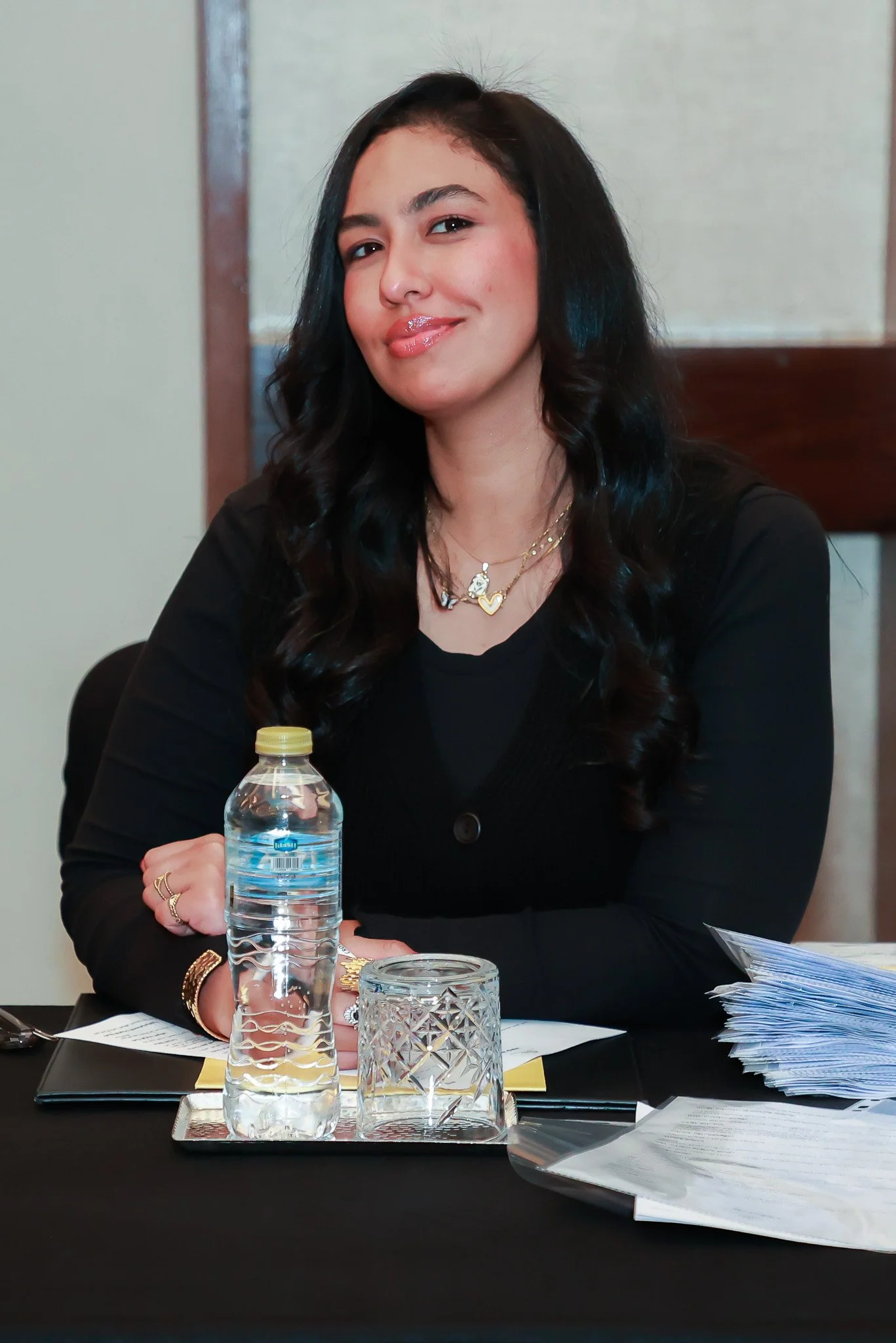 A woman with long black hair, wearing jewelry, sitting at a table with a water bottle, glass, and papers.
