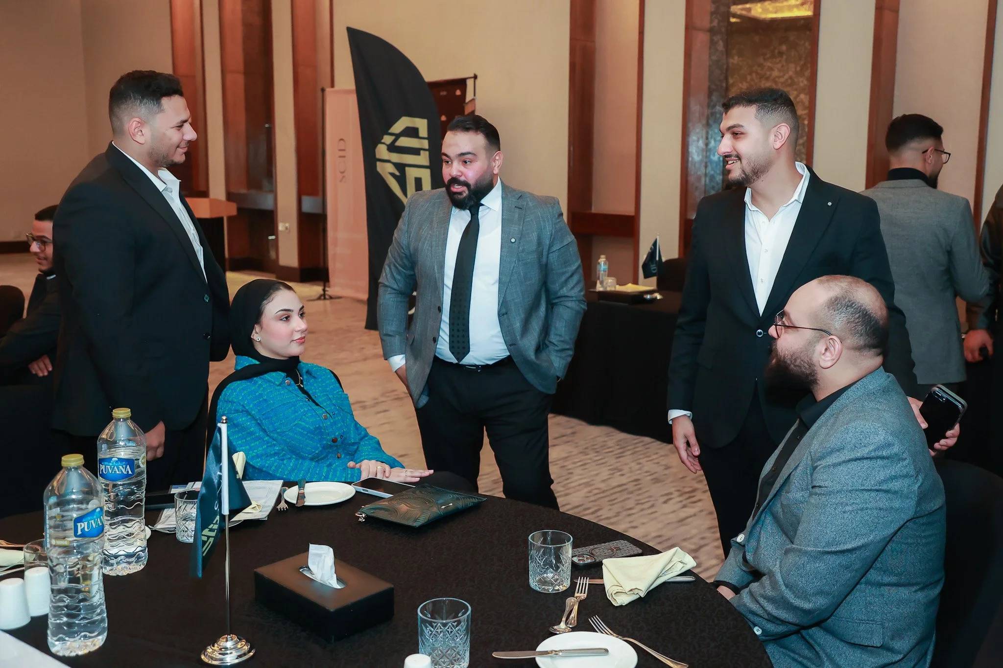 Group of five people, four men and one woman, engaged in conversation around a table at a formal event or conference. The woman is seated with a black headscarf, and water bottles are on the table along with dining utensils and napkins.