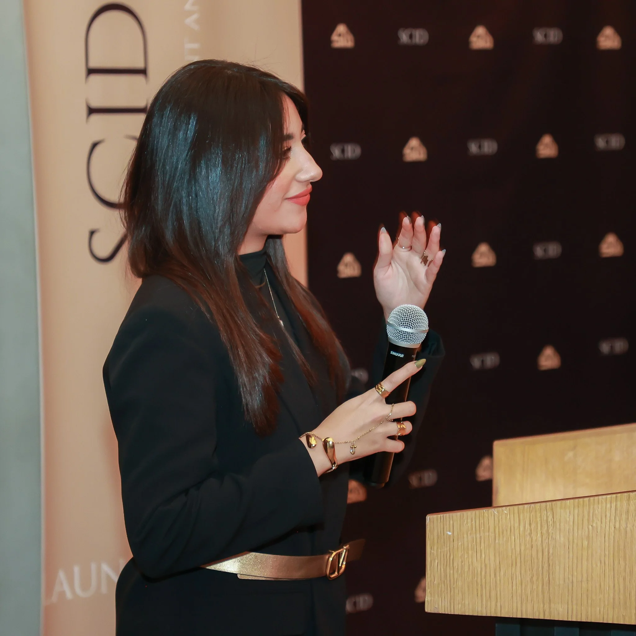 A woman with long dark hair, dressed in black with gold jewelry, holds a microphone and gestures with her right hand at a podium during an event.
