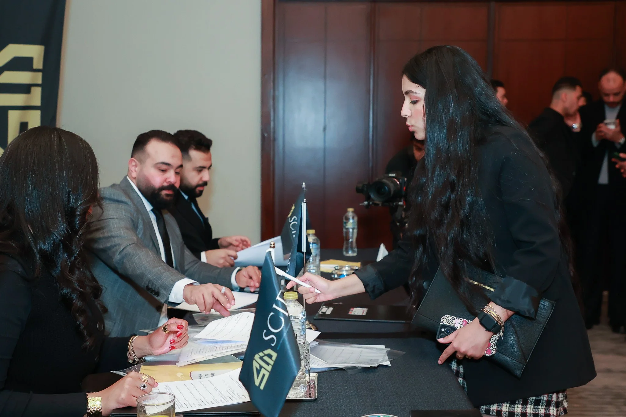 A woman at a conference or registration table is handing a document or registration form to a man seated at the table, with other seated adults engaged with documents and laptops, and a backdrop with a partial logo and wooden paneling.