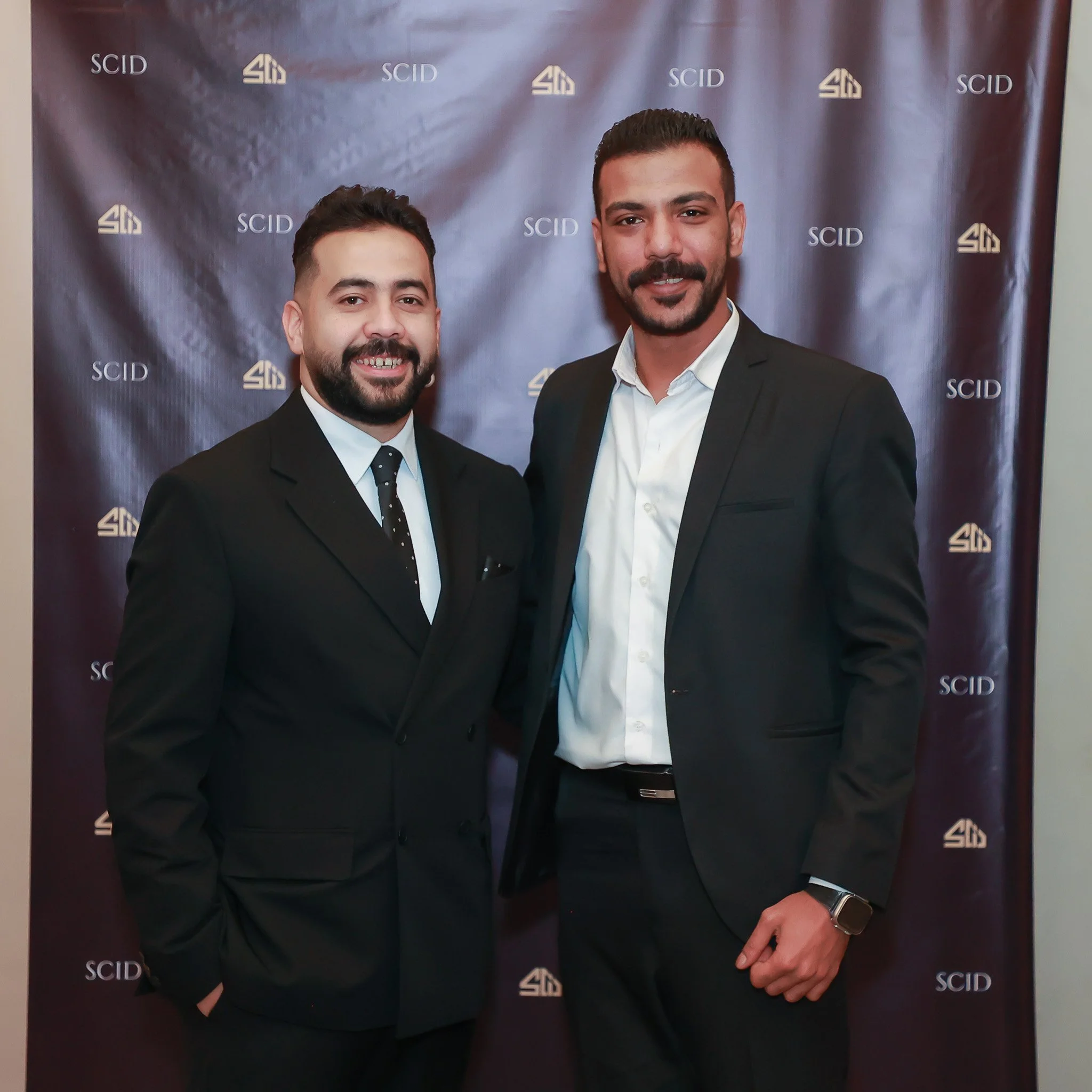 Two men in black suits, white shirts, and black tie, smiling and standing close together in front of a dark backdrop with the logo 'SCID' repeated across it.