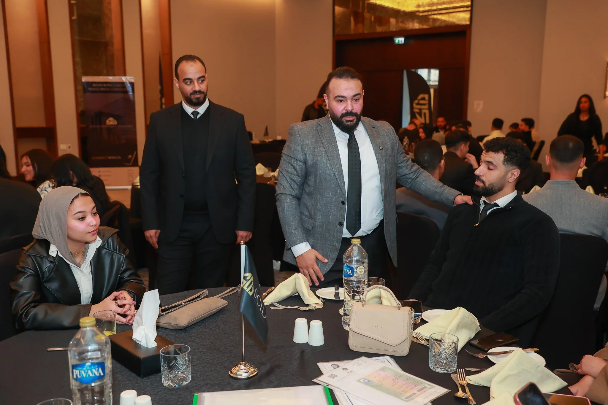 A formal gathering with several people seated at round tables. A man in a grey suit is speaking to a seated man dressed in black, while another man in a black suit watches. A woman in a grey hijab sits at the table, looking ahead. The table is set wi