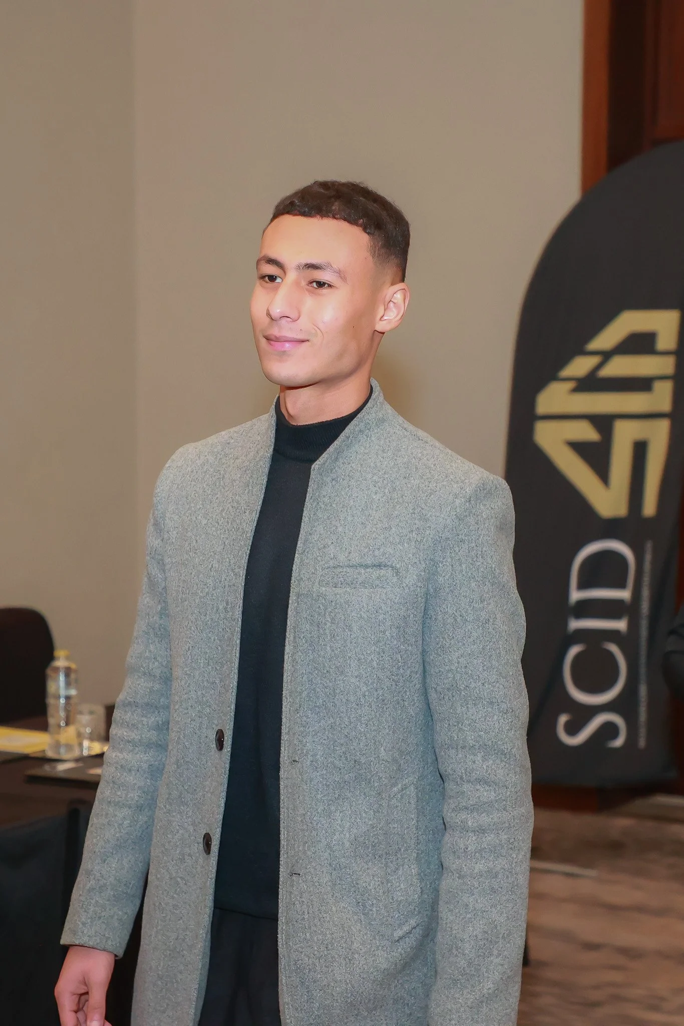 A young man with short dark hair and light skin is wearing a grey coat over a black turtleneck, standing indoors during a formal event.