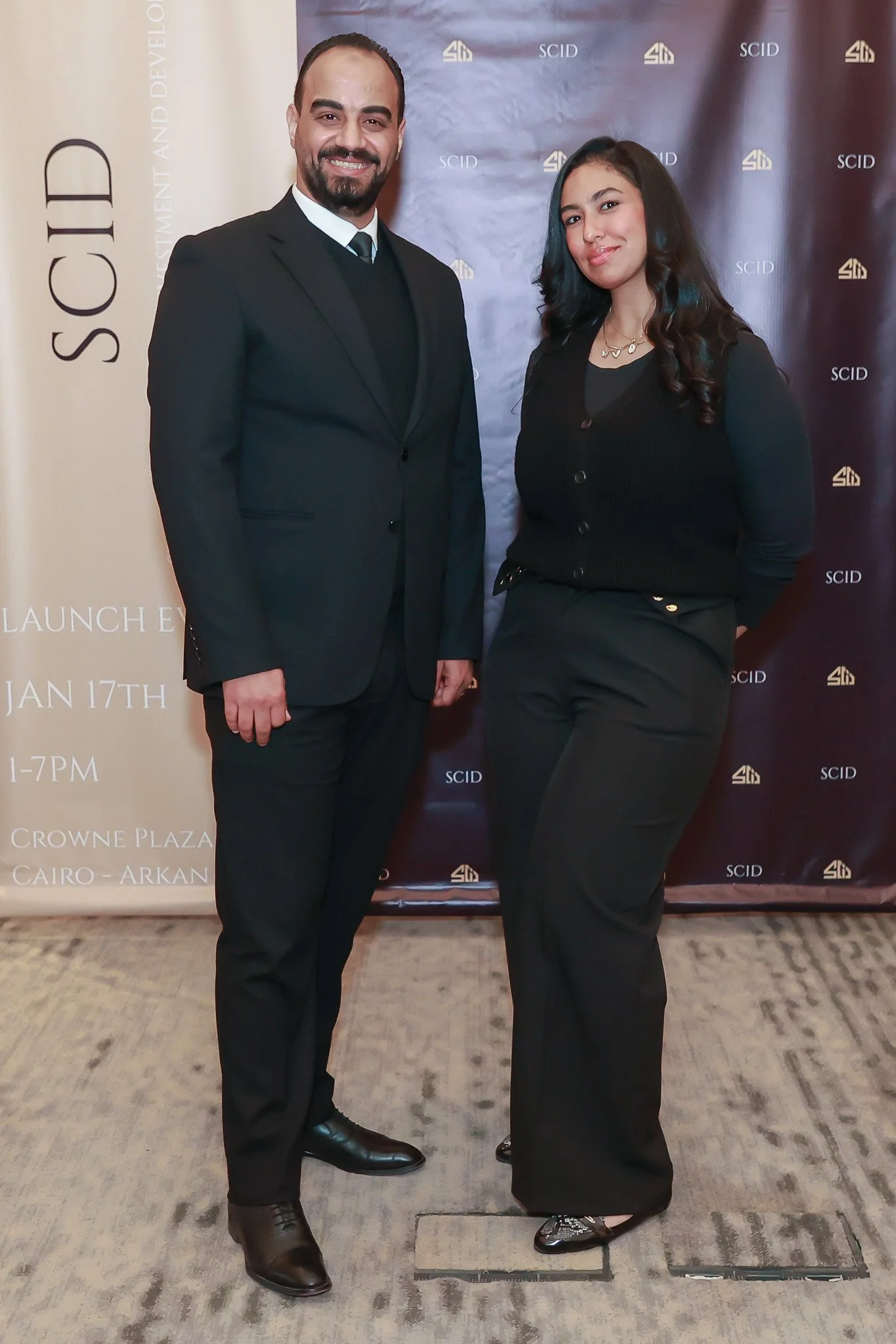 A man and woman standing side by side in formal attire, smiling, at an event with a backdrop featuring logos and information.