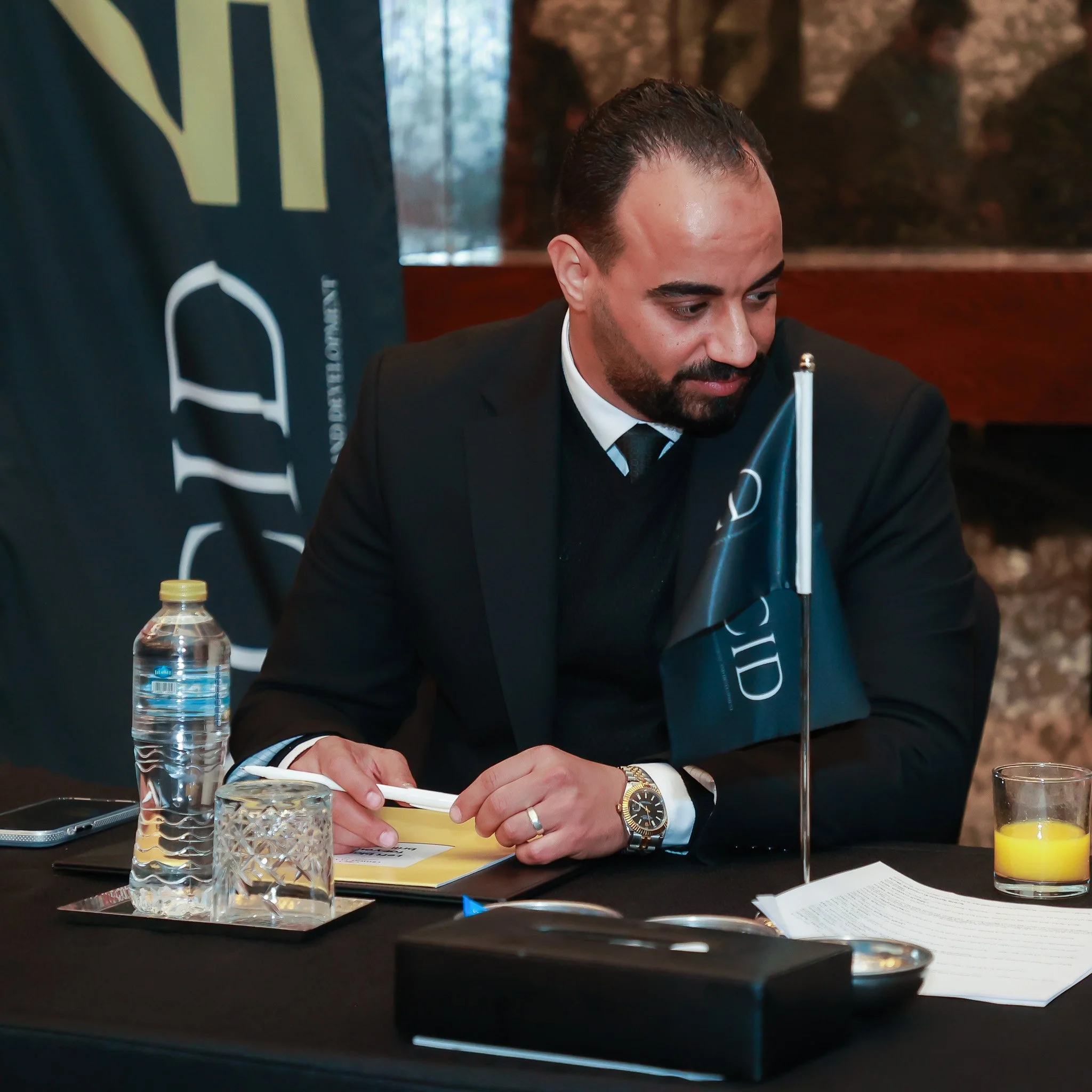 A man in a black suit sitting at a conference table with a small flag in front of him, a water bottle, a glass of orange juice, and papers.
