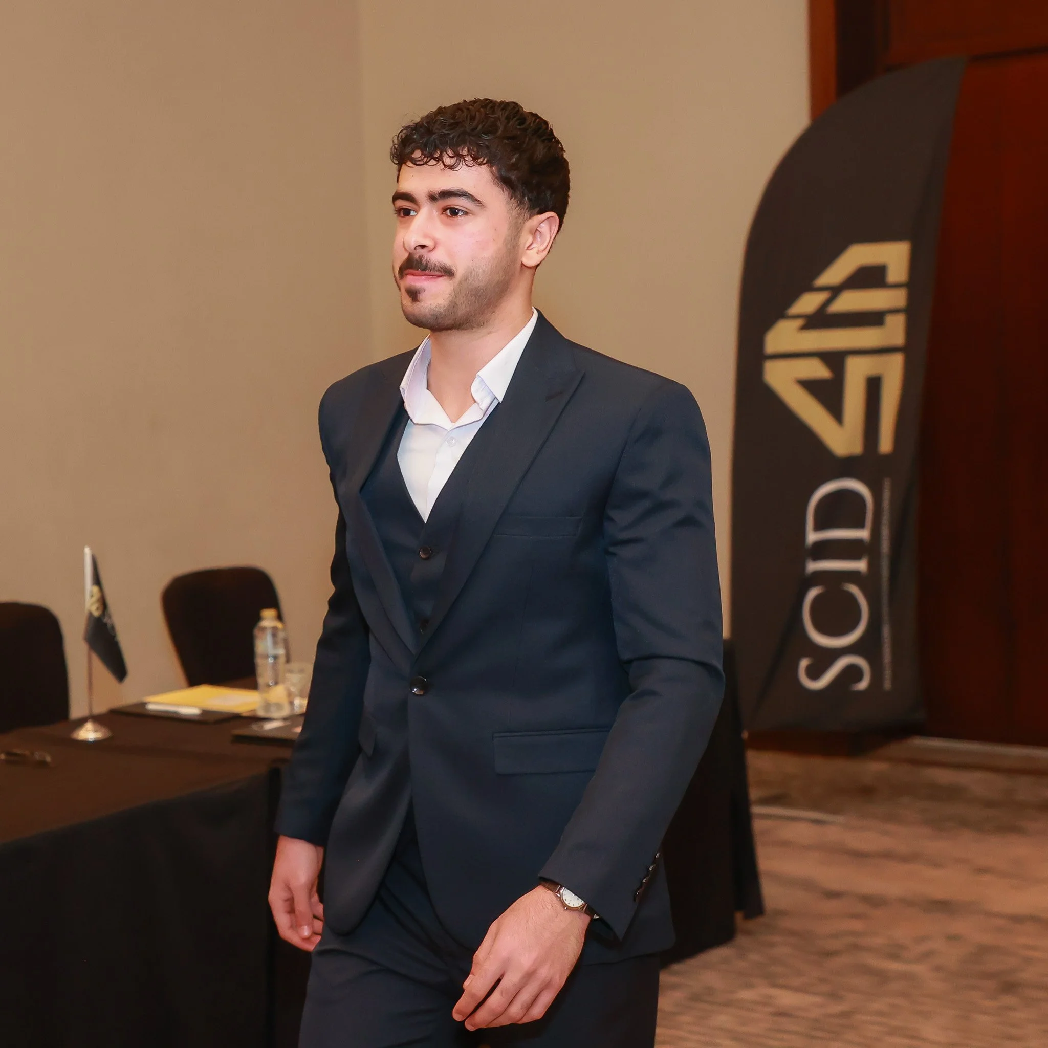 A man in a dark suit with a white shirt stands in a conference room, with a S2ID sign on a flag in the background.